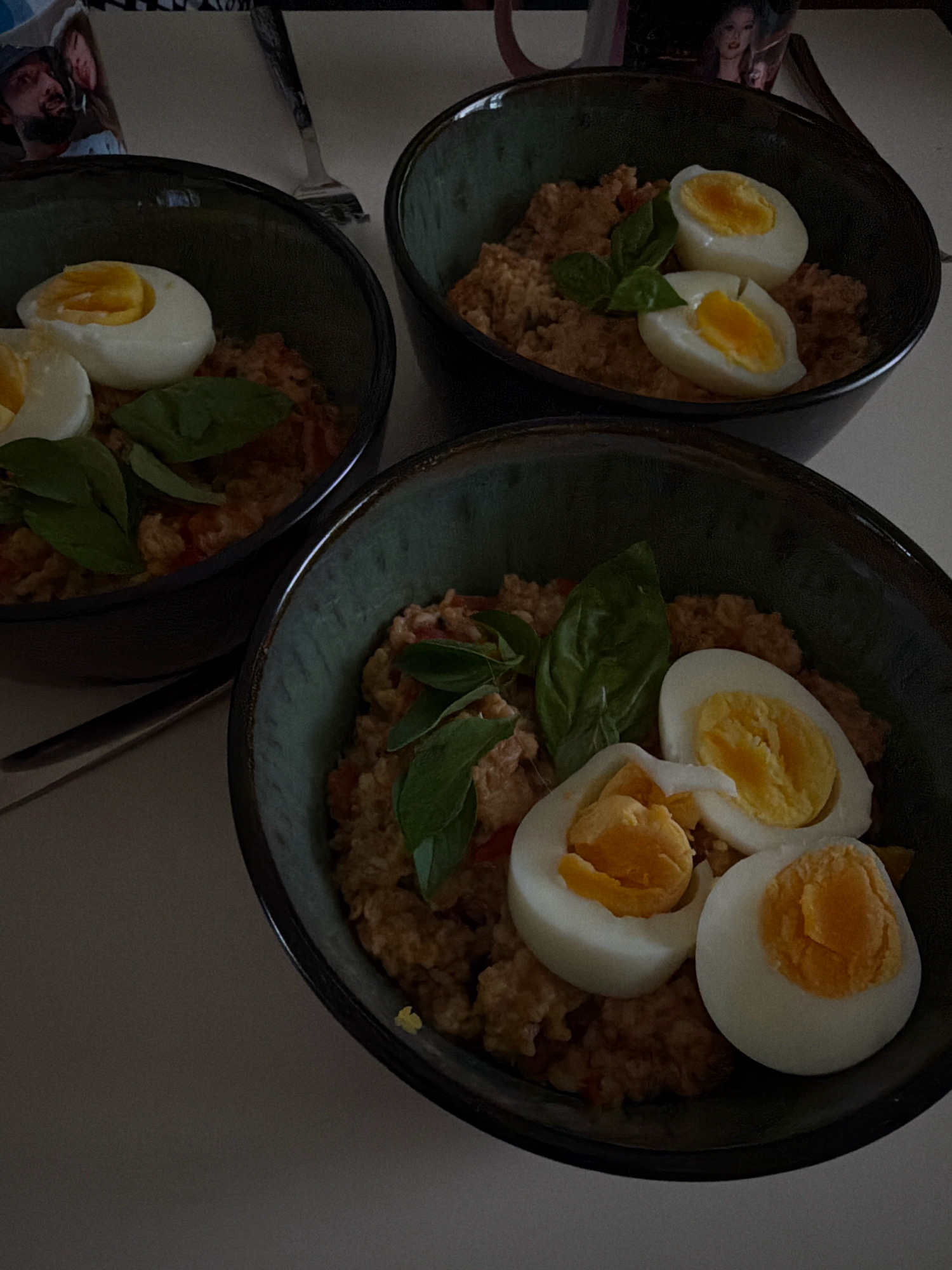 The image shows three bowls of food, each topped with sliced hard-boiled eggs and garnished with basil leaves. The bowls contain a textured, brownish mixture that appears to be a savory porridge or rice dish with visible red flecks, possibly tomatoes. Two of the bowls are in the foreground, with the third slightly further back. The setting appears to be indoors on a light-colored surface. In the upper left corner, a photograph or print is visible, showing two individuals. The lighting is dim, creating shadows. There is no indication of the specific location in Sharm El Sheikh, Egypt.