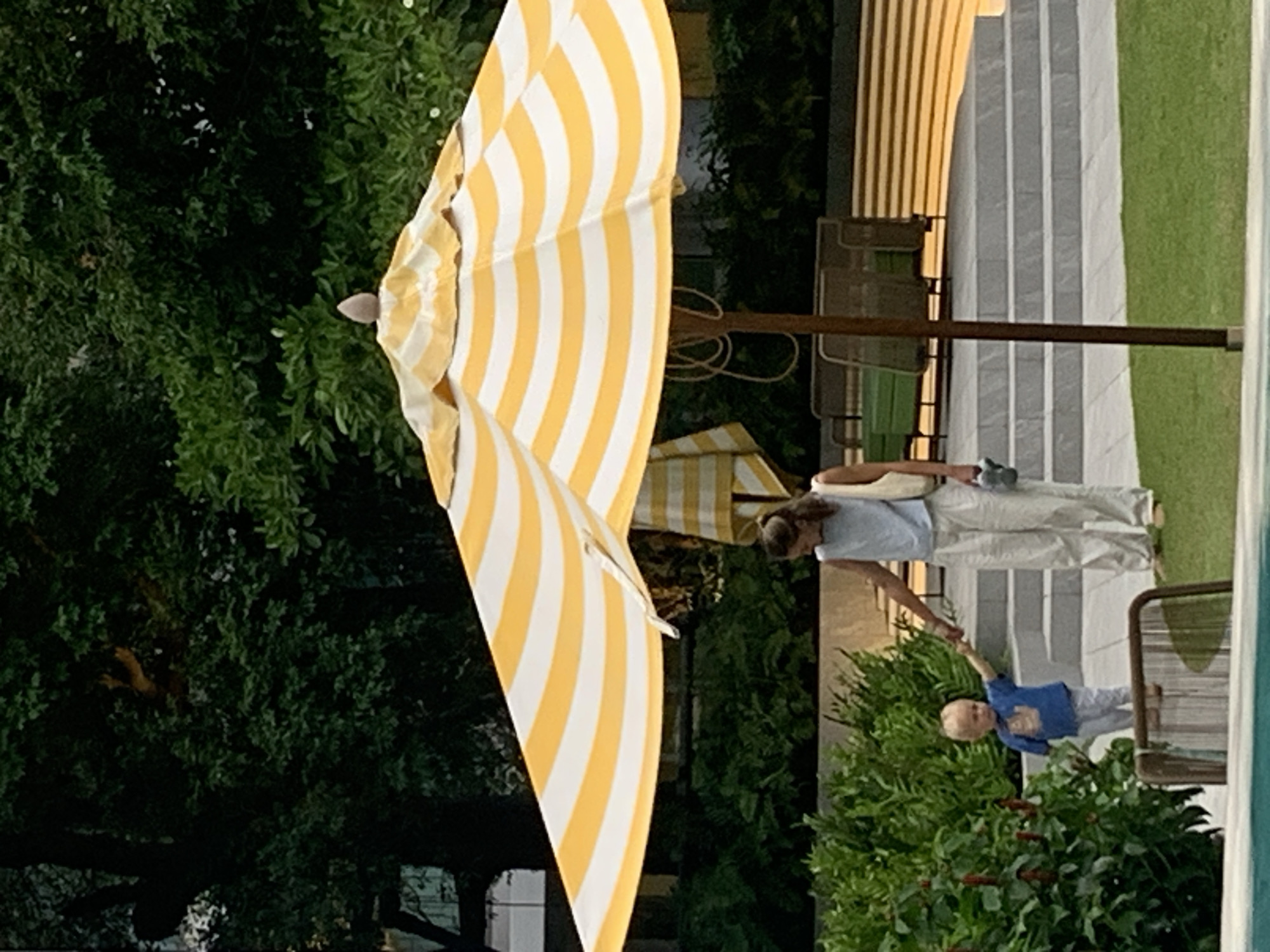 The image depicts an outdoor scene with two people and a large, partially unfurled yellow and white striped umbrella. A woman, wearing light-colored pants and a blue shirt, is interacting with a young child. The child, dressed in a blue shirt and light-colored pants, is reaching out a hand, possibly being held by the woman or about to be led. They are standing near lush green foliage and a paved area with a checkered pattern. In the background, there is more greenery, a structure with vertical yellow and white stripes, and what appears to be a poolside area, suggested by a sliver of turquoise water visible on the right edge of the frame. The scene appears to be set during the daytime, with the lighting suggesting a sunny day. There is no visible text in the image. The overall impression is one of a relaxed outdoor gathering or outing.