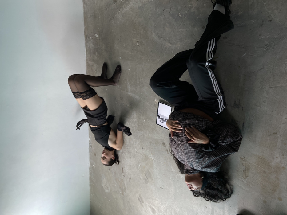 The image depicts an indoor scene, presumed to be in the city of London, United Kingdom, within a minimalist room featuring a plain light-colored wall and a textured concrete floor. Two individuals, identified as adult females, are present, both lying on the floor.
The first individual, positioned towards the upper portion of the frame, is lying supine with her back arched, hips elevated in a bridge-like pose, knees bent, and feet flat on the floor. Her arms are bent, with hands raised near her head, fingers splayed. She is dressed in a black sleeveless crop top, black shorts, black sheer thigh-high stockings with lace trim, and black sheer gloves. Her dark hair is pulled back.
The second individual, positioned in the foreground, lies supine with her head to the left, eyes directed upwards. Her legs are bent and splayed to her right. Her left arm is positioned across her chest, with her right hand resting on her abdomen. She is wearing a plaid long-sleeved shirt, black athletic track pants with three white stripes along the sides, a black headband, and dark athletic shoes. Her long, dark, curly hair is spread around her head on the floor.
A white tablet computer rests on the concrete floor between the two individuals, slightly to the right of the second individual's head, displaying a black-and-white image on its active screen. There is no direct physical interaction between the two individuals.