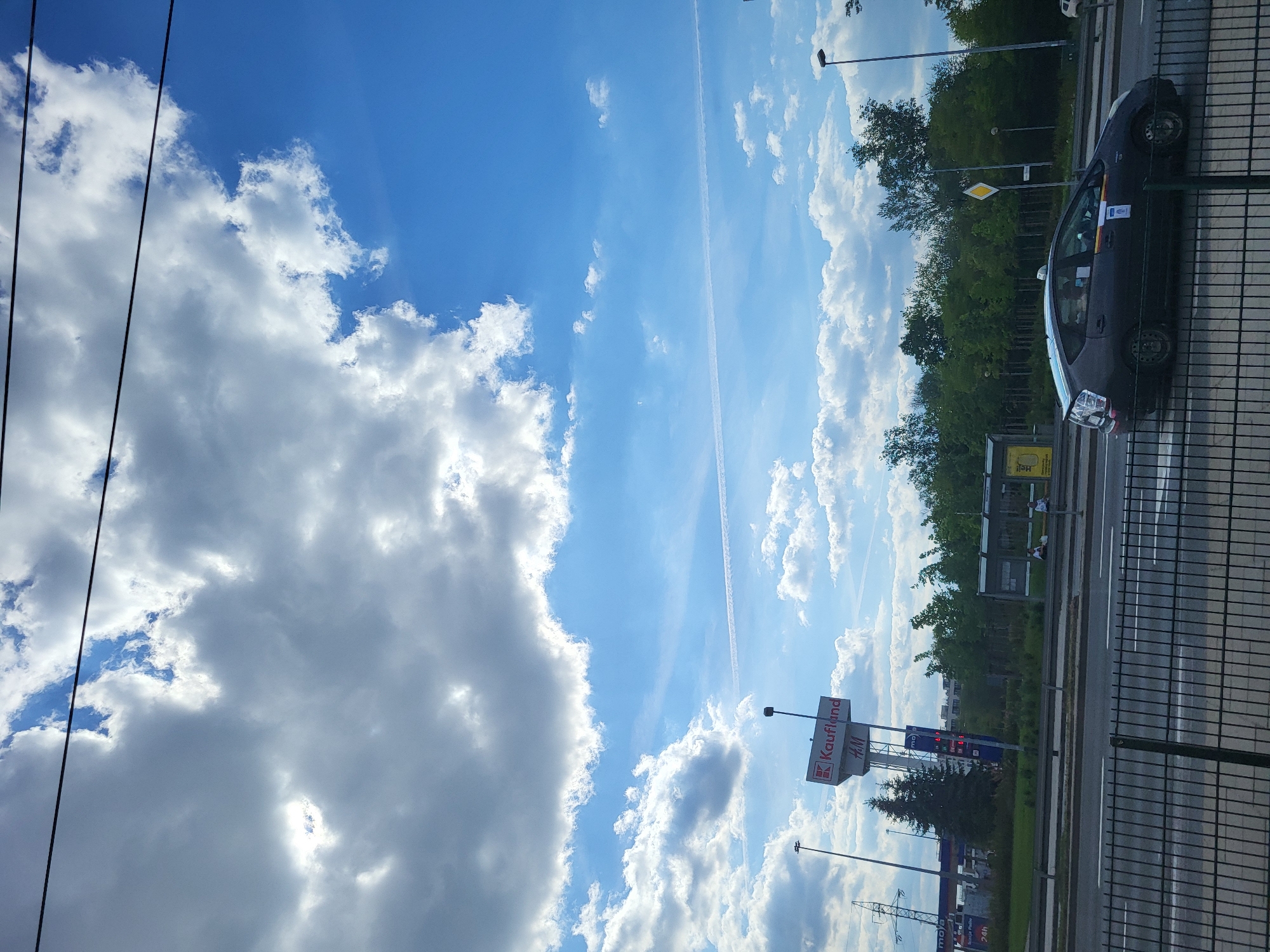 The image shows a partly cloudy blue sky and a view of a city street. In the foreground is a black metal fence. To the right is a black car parked next to the street. Further into the scene are trees lining the street. A sign for Kaufland and an H&M are visible. The location is not specified.