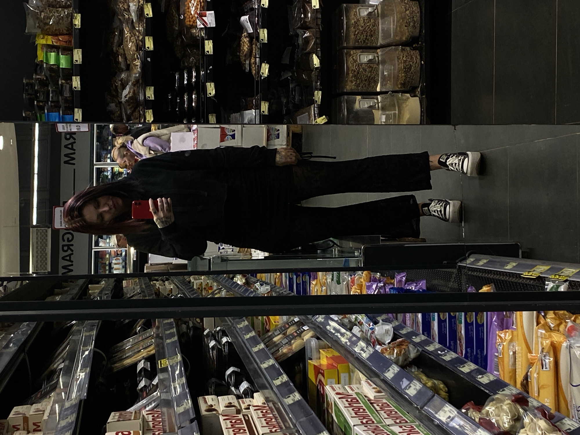 An image, captured in a grocery store or supermarket aisle in Ulcinj, Montenegro, shows a young person taking a mirror selfie. The subject, positioned centrally, is reflected in the polished dark floor, and appears to be standing upright against a backdrop of retail shelving. She wears dark long-sleeved clothing, dark trousers, and black-and-white sneakers, possibly Converse, with white laces. Tattoos are visible on her left hand and wrist as she holds up a red smartphone, which obscures most of her face. Her long, dark reddish hair cascades over her shoulder.
In the background, on the left, vertical shelves hold clear containers with various bulk goods like nuts, seeds, and dried fruits, alongside packaged items in bags. To the right, horizontal shelves display rows of packaged foodstuffs, including cereals, snacks, and boxes, some with "SKD" and "76V" visible on their labels. Another person with blonde hair, possibly an employee or another shopper, is partially visible and reflected in the background. The store environment is illuminated by artificial lighting. On the reflective floor, the Cyrillic word "МАЯ" (MAY) is visible as a reflection from an overhead sign. The overall scene suggests a casual shopping experience in a modern retail setting.