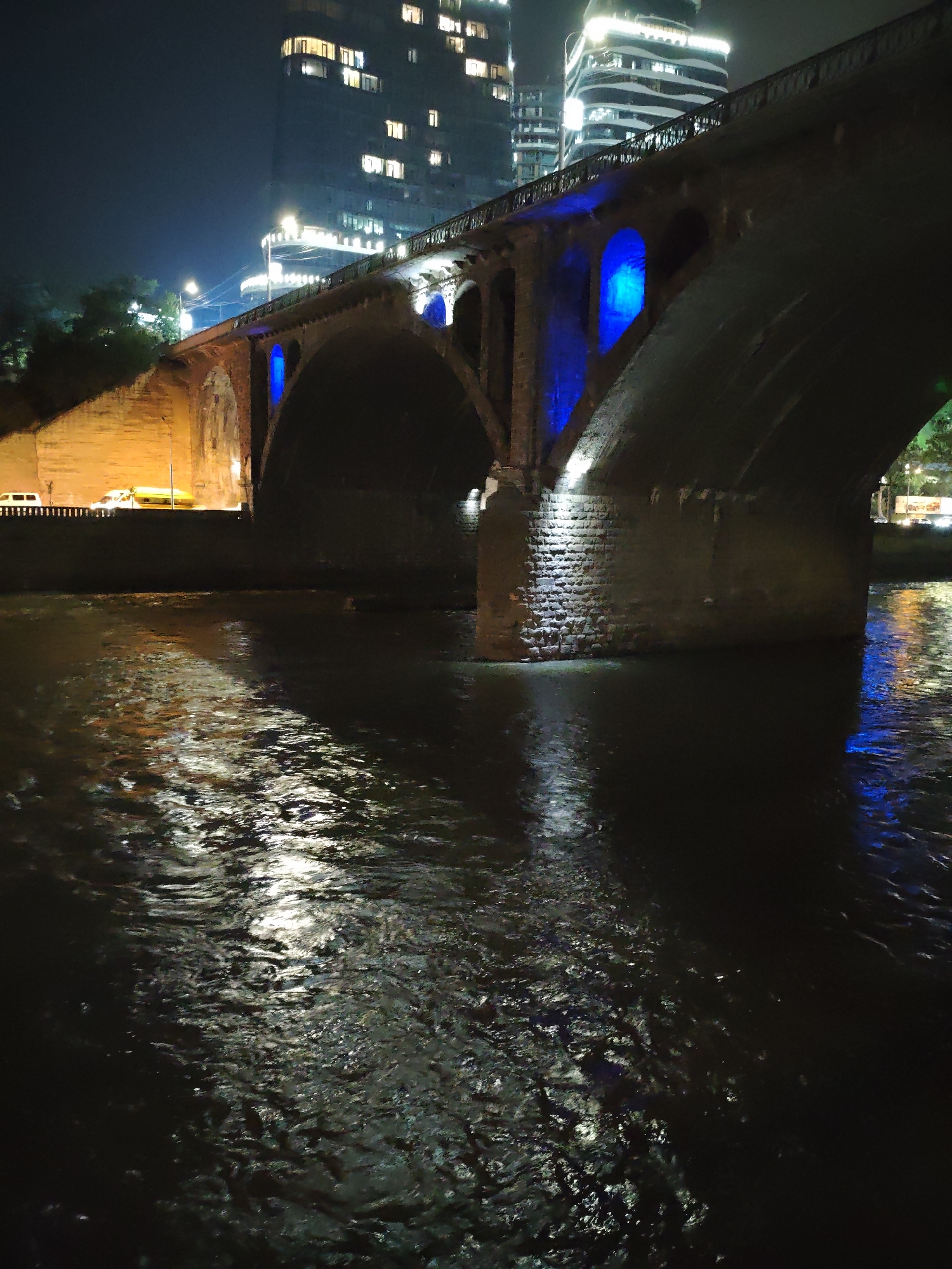 The image depicts the Peace Bridge in Tbilisi, Georgia, at night. The bridge is illuminated with blue lights, and its reflection can be seen on the surface of the Kura River below. Modern buildings with illuminated windows are visible in the background. The scene is characteristic of the urban landscape of Tbilisi at night, showcasing a blend of historic and contemporary architecture. The Kura River flows through the city, serving as a central element in the urban scenery.