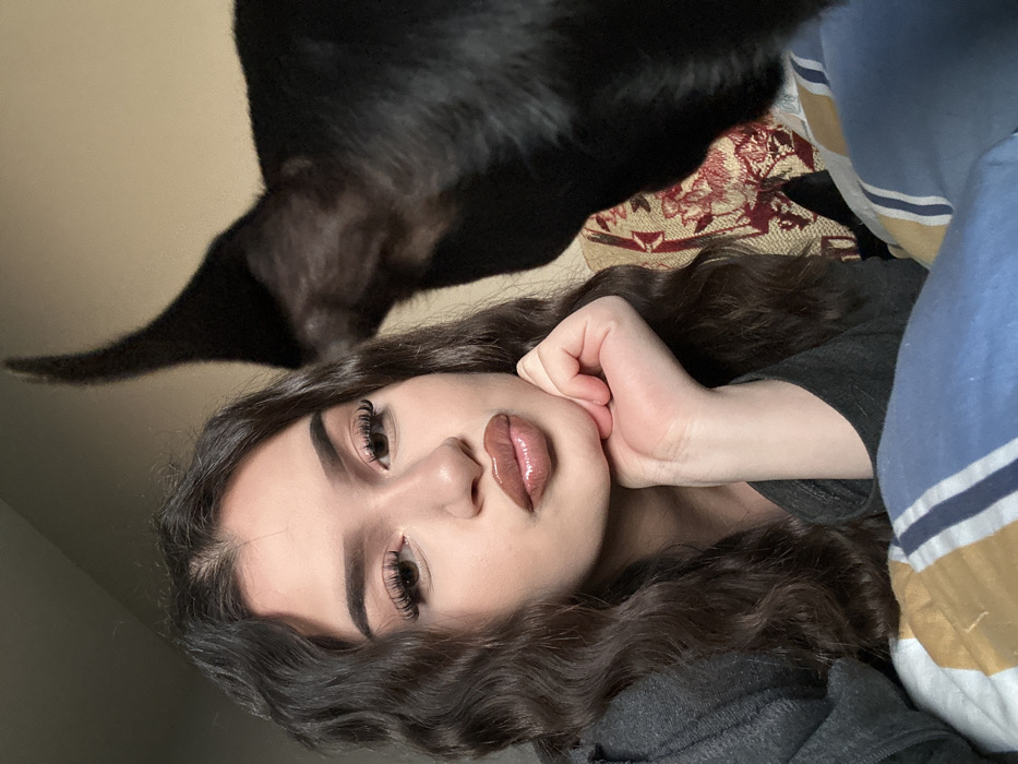 An indoor scene in Okop, Bulgaria, features a female subject and a dog. The female, centered on the left, is an adult with light skin, dark wavy hair, and visible makeup including enhanced eyelashes and glossy, pouted lips. She wears a dark grey long-sleeved garment and rests her right hand, fingers curled, beneath her chin. Her gaze is directed towards the camera. To her right, partially visible, is a solid black, short-haired dog, showing the right side of its head, including a distinctly pointed ear. The background includes a plain, light-colored wall at the top and a patterned textile, possibly a pillow or blanket, with red, gold, and cream intricate designs. The foreground shows blue and beige striped bedding or blankets.