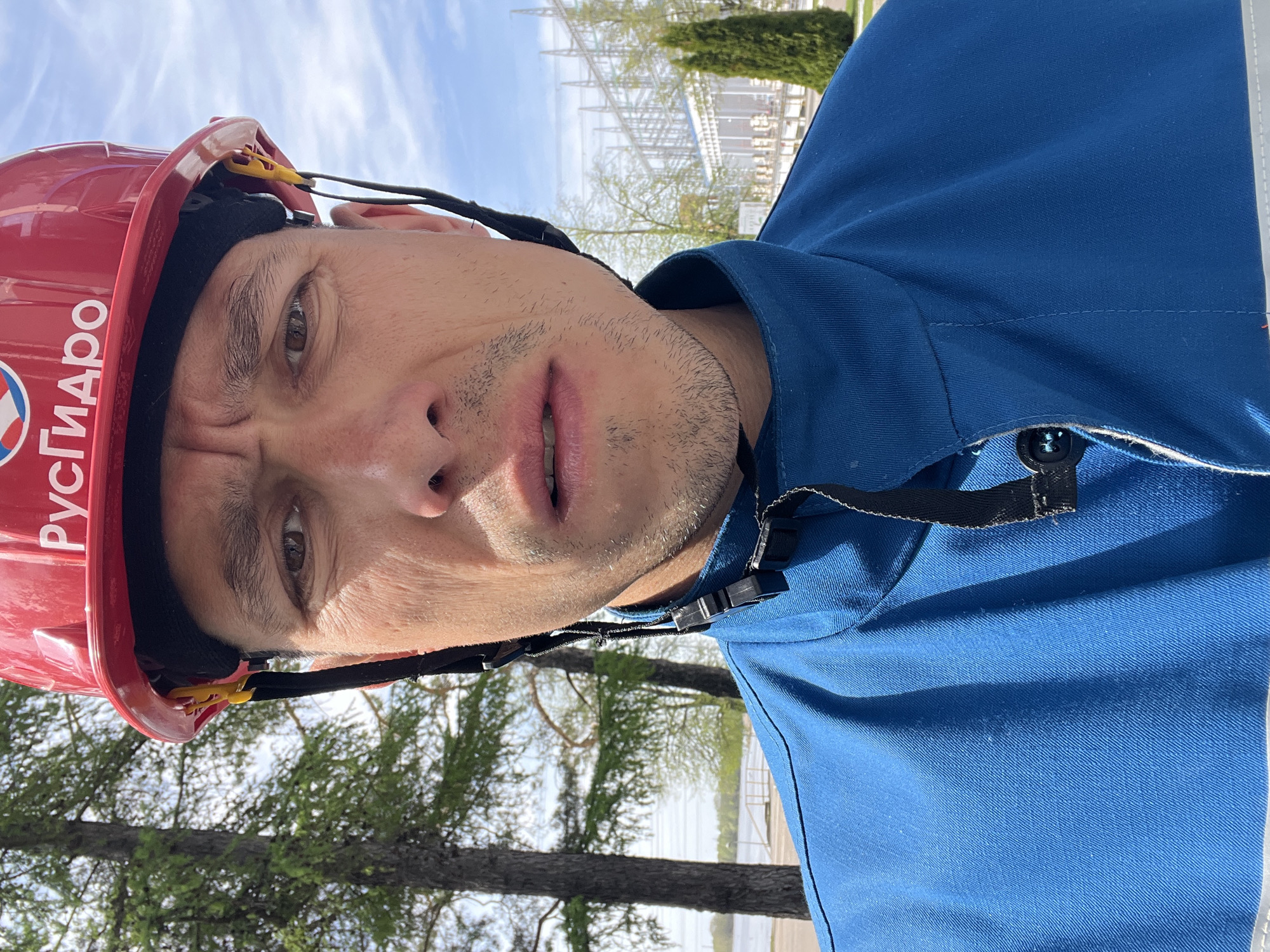 A man is looking at the camera. He is wearing a red hard hat with the word "РусГидро" printed on it. He is also wearing a blue work shirt. The background shows trees, water, and structures that suggest a power plant. The location is likely Chaikovsky, Russia, given the hard hat's inscription likely referencing RusHydro and power plant structures.