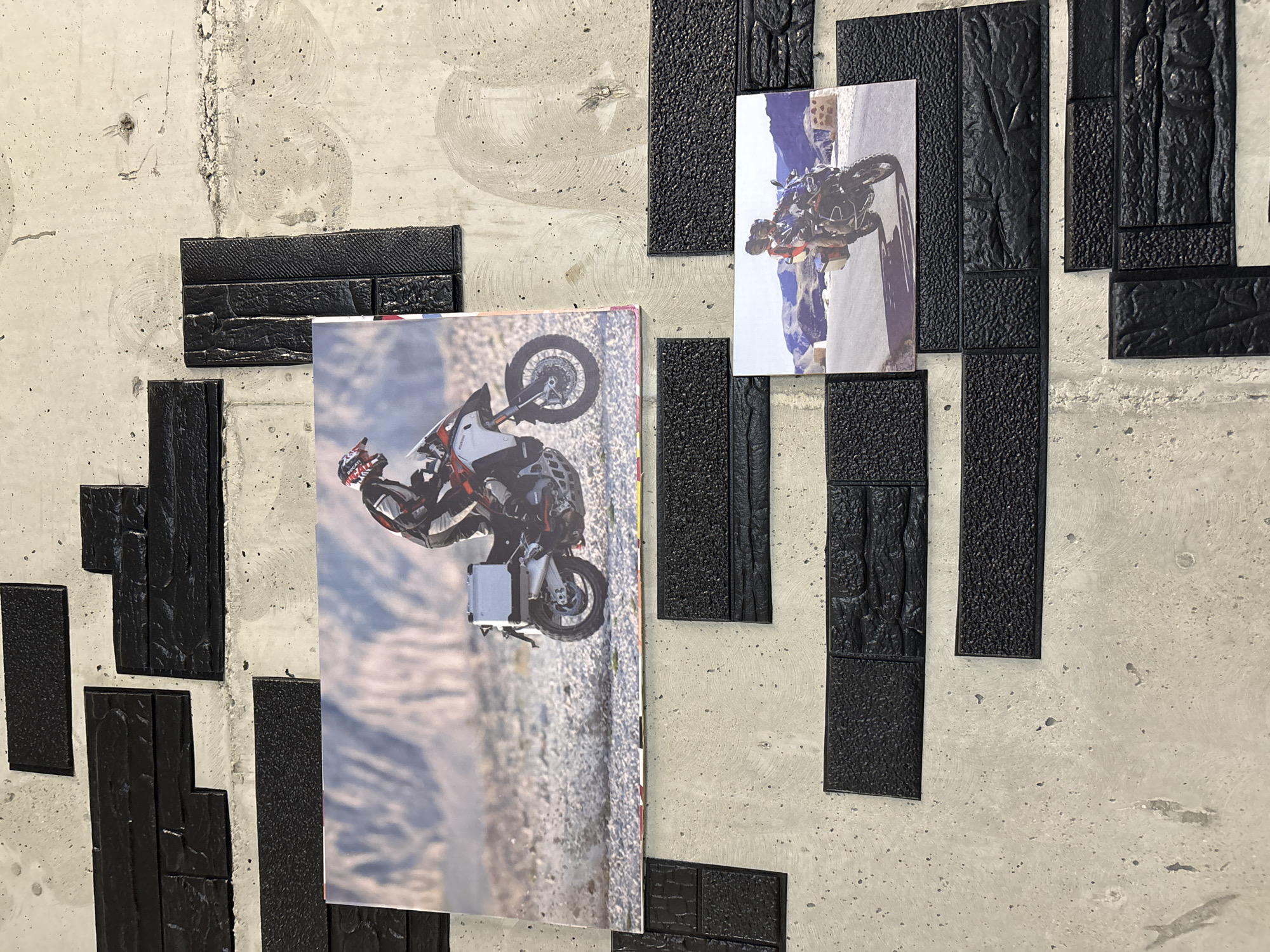 This is a photograph of a textured concrete wall in Tbilisi, Georgia, adorned with two framed pictures of motorcycles and their riders. The pictures are placed on the wall, which has a rough, speckled gray concrete surface. Black, rectangular, textured elements resembling bricks or tiles are adhered to the wall in a staggered pattern, creating a decorative border around the photographs.
The top picture, larger than the bottom one, shows a person in full riding gear on a white and red adventure motorcycle. The motorcycle is positioned on a gravelly, off-road terrain with a blurred background of arid hills. The rider is wearing a helmet and appears to be in motion.
The bottom picture, smaller and positioned below the first, depicts two people on a dark-colored adventure motorcycle, riding on a paved road that curves through a mountainous landscape. The sky is blue, and the scene suggests a scenic ride.
The overall impression is that of an indoor space, possibly a home, garage, or commercial establishment, decorated with motorcycle-themed imagery. The black textured elements create a stark contrast against the concrete wall and the photographs, adding a graphic and modern aesthetic to the display.