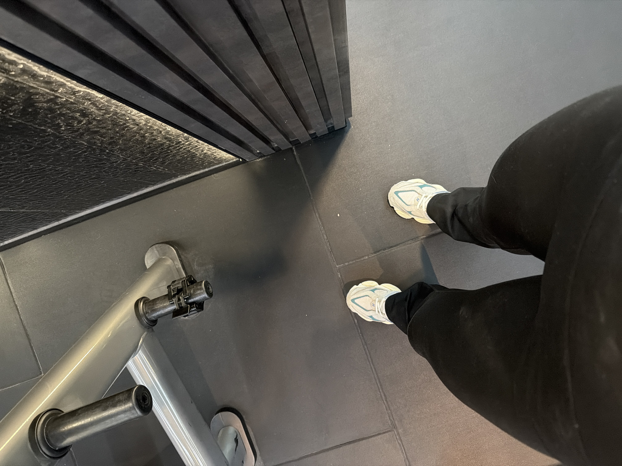 A first-person perspective photograph taken indoors in Plovdiv, Bulgaria, shows a person wearing black leggings and off-white sneakers with light blue accents. The person is standing on a dark gray tiled floor adjacent to a metallic gym machine with chrome and black elements. Part of a dark-colored wall with a textured surface is seen along the top left edge of the frame.
