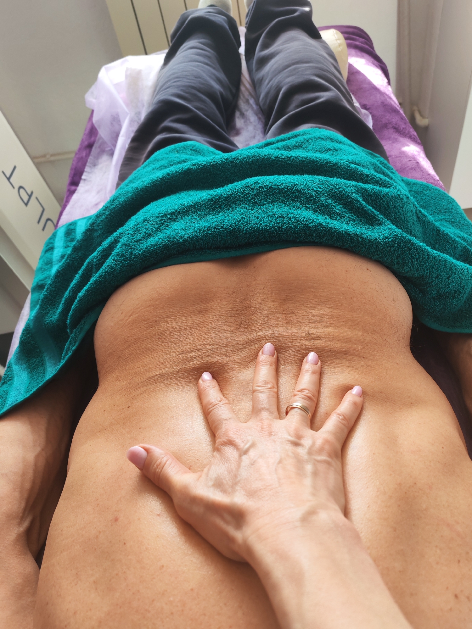 A person lies supine on a bed, with their black-clad legs extending upward. A turquoise towel covers their lower back. A hand is placed on the bare skin of their upper back, fingers spread. The person's nails are painted a light pink. The background consists of a light purple sheet and what appears to be a hospital or clinic interior. There's no information that would allow one to say this scene is specifically in Chaikovsky, Russia.