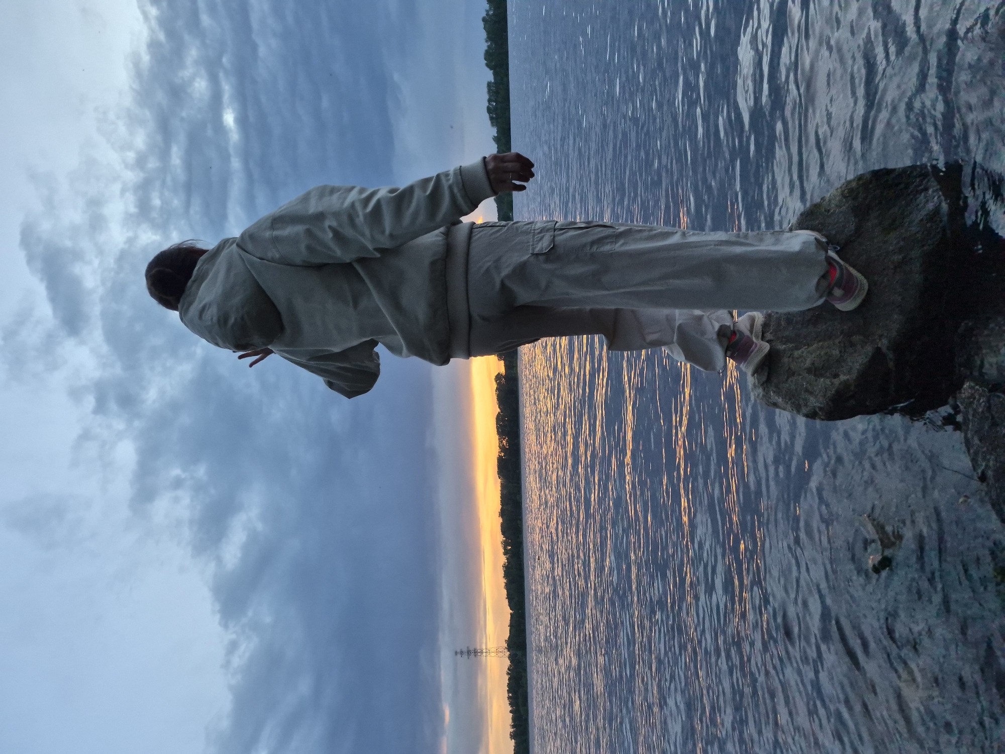A person in a light-colored jacket and pants stands on a rock by the water's edge, gazing towards the water. The sky above is filled with clouds, and the water reflects the sunset. A power line is visible in the distance. The location is Unknown, Unknown.