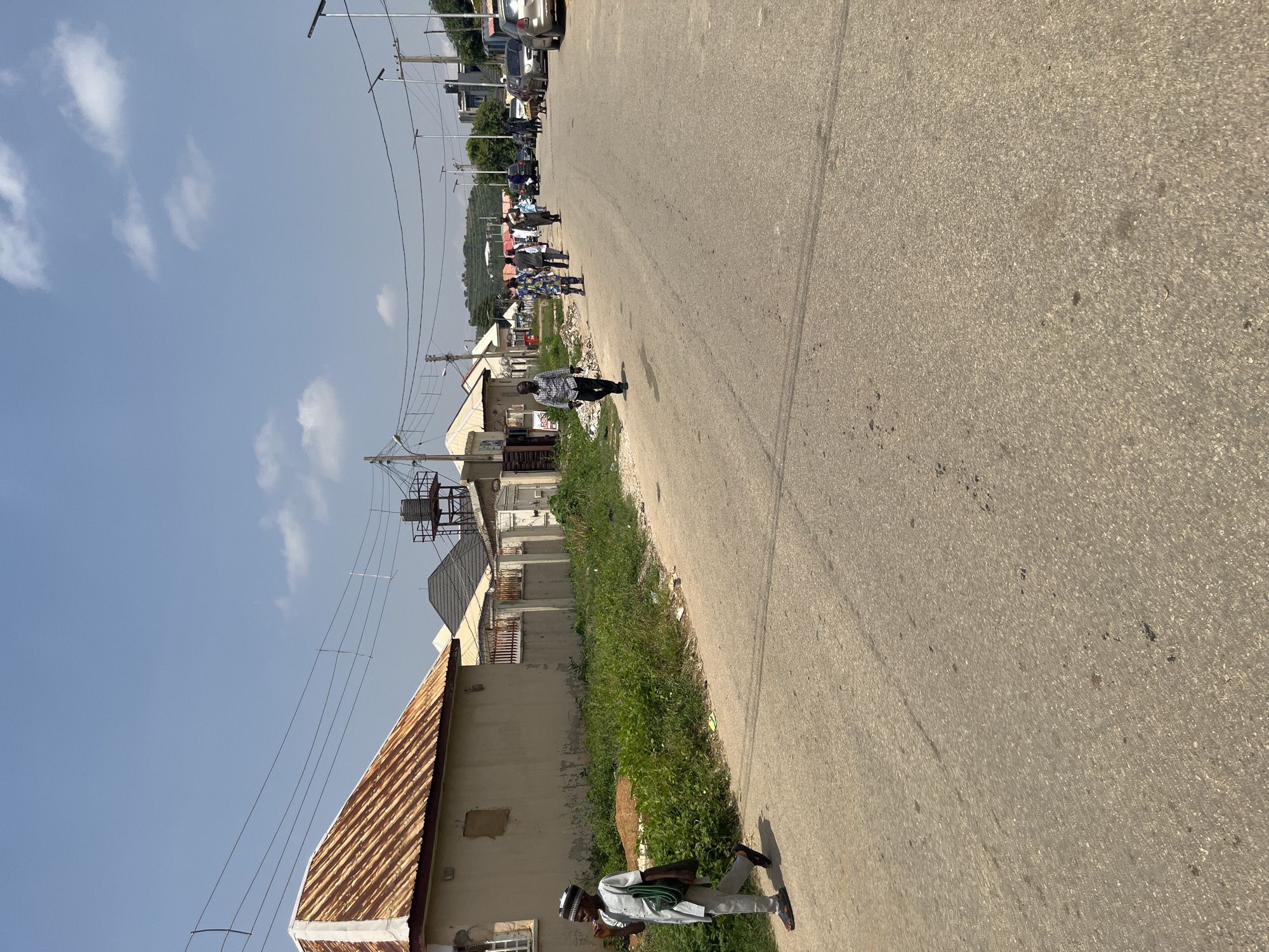 This is a medium shot of a street scene in Abuja, Nigeria, captured on a bright, sunny day, indicated by the clear sky with scattered clouds and strong shadows. The perspective is slightly tilted, giving a dynamic feel to the image.
The primary subjects are several people walking along a paved road. In the foreground, a man in traditional Nigerian attire, a white kaftan and a patterned cap, walks towards the right of the frame. He has a dark bag slung over his shoulder. Further down the road, a group of people are walking in the same direction, some dressed in casual wear, others in what appears to be uniforms or traditional clothing. They are spread out along the road and the grassy verge.
The setting is a suburban or semi-urban street lined with buildings. On the left side of the road, there are houses with corrugated iron roofs and beige-colored walls. Some buildings have visible windows and architectural details. Power lines and poles are prominent throughout the scene, indicating infrastructure. In the background, a gentle slope covered with trees suggests a hilly or elevated area. Cars are visible parked on the side of the road further down, and some traffic can be seen in the distance.
The overall atmosphere appears to be one of everyday life, with people going about their business on a clear day. There is no visible text in the image. The time of day is likely midday, given the intensity of the sunlight and the length of the shadows.