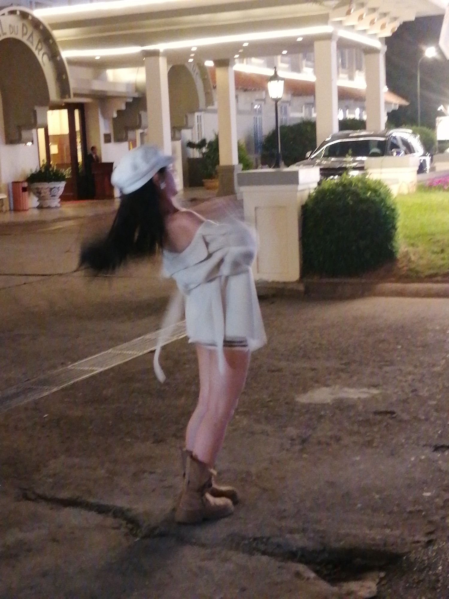 The image depicts a young woman in a casual outfit, including a white cap and beige boots, standing on a paved area outdoors. She appears to be mid-motion, possibly turning or dancing, with her dark hair blurred from movement. She is wearing a light-colored dress or top with flowing sleeves and a skirt. The setting seems to be in front of a building with arched entrances and outdoor lighting, suggesting it might be a hotel or a similar establishment.
To the right of the woman, there is a large, neatly trimmed green bush and a patch of grass. Behind her, a dark-colored SUV is parked. The overall lighting suggests it is evening or night, with artificial lights illuminating the scene. The ground is dark and appears wet in some areas, possibly indicating recent rain or just dampness from the evening.
The building in the background has a classical architectural style with pillars and arches. Some text is visible on the building's facade above an arched entrance, reading "LE DU PARC". The atmosphere seems relaxed and possibly festive, given the woman's dynamic pose. The location is identified as Da Lat, Vietnam.