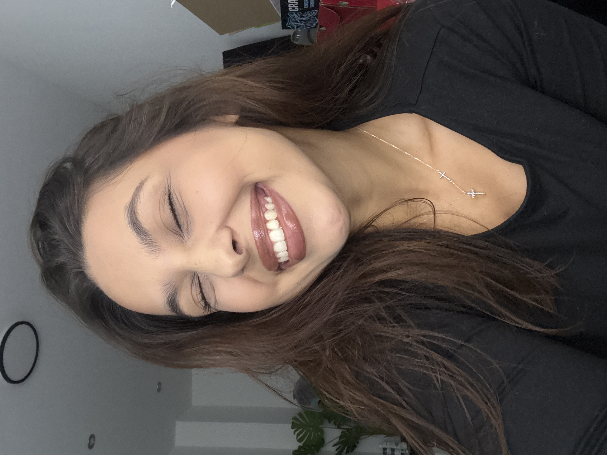 A young woman with long, dark brown hair is captured in a close-up, joyful moment, with the image rotated 90 degrees clockwise. She has fair skin and is smiling broadly, her eyes closed in an expression of evident happiness or laughter. Her full lips are adorned with glossy, brownish-pink lipstick, and her white teeth are visible as she grins. She is wearing a plain black long-sleeved top. A delicate gold-colored chain necklace with two small cross pendants, one simple and the other sparkling with what appears to be tiny crystals, rests on her collarbone.
The background indicates an indoor setting. Behind her head, a light-colored wall displays a black circular decorative object. To the upper left of the frame (due to the image's rotation), a dark object or box is partially visible, featuring white text that reads "CRAN" and "ENERGY DRINK." Further in the background, hints of green foliage, possibly from a large houseplant, and other indistinct objects suggest a typical room interior. The lighting is soft, highlighting the woman's expressive face and the cheerful emotion conveyed.