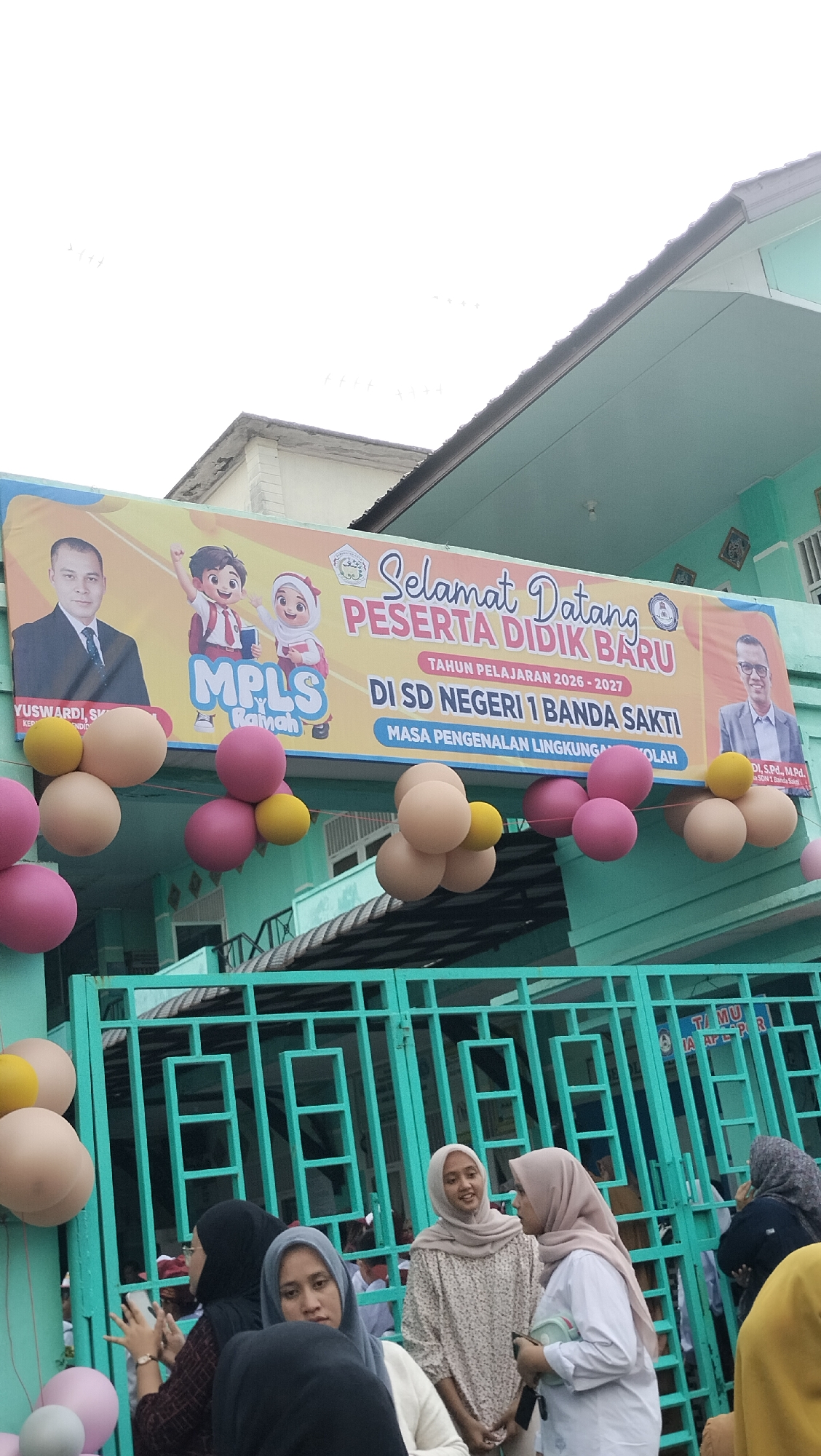 The image shows a banner in front of a school in Lhokseumawe, Indonesia, that reads "SELAMAT DATANG PESERTA DIDIK BARU TAHUN PELAJARAN 2026-2027 DI SD NEGERI 1 BANDA SAKTI MASA PENGENALAN LINGKUNGAN SEKOLAH". The banner features cartoon children and portraits of two men, presumably school staff. Balloons in pink, yellow, and beige are hung in front of the banner and the school entrance. A green metal gate with decorative elements is visible in the foreground. Several people, mostly women wearing hijabs, are gathered in front of the gate. The building appears to be painted in light green and white.