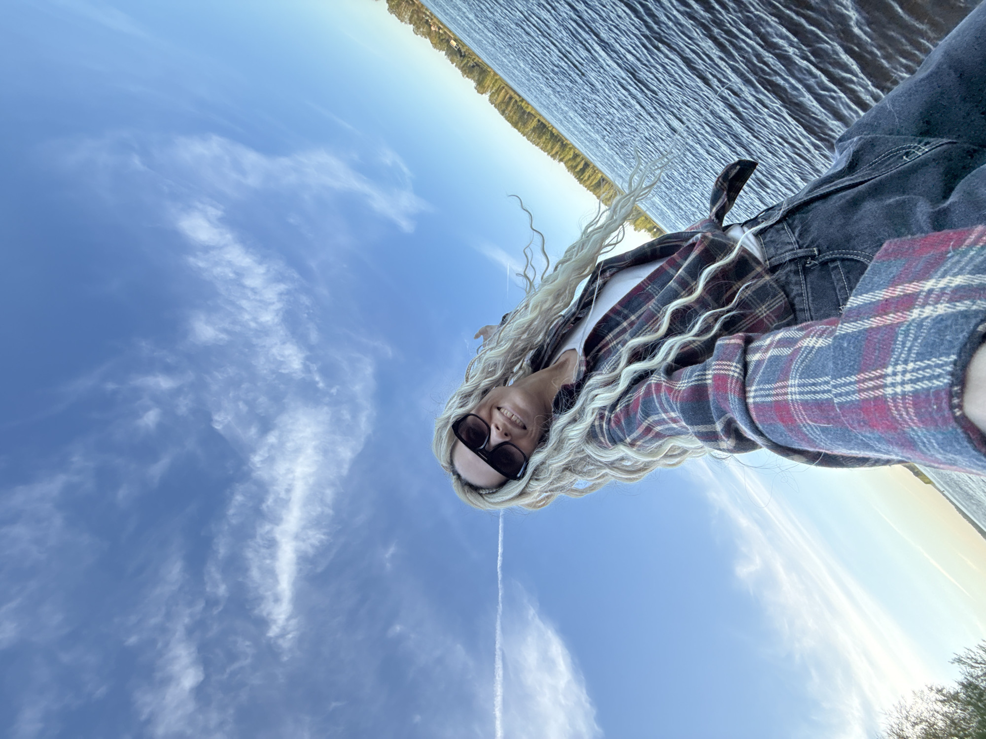 A woman wearing dark sunglasses, a white shirt, and a plaid shirt is standing outside on a sunny day with a body of water visible in the background. The woman is wearing dark colored jeans. She has long blonde braided hair. The body of water has trees visible along the shore. The sky is blue and has wispy clouds in it. Rusanovo, Russia is not identifiable in this photo.