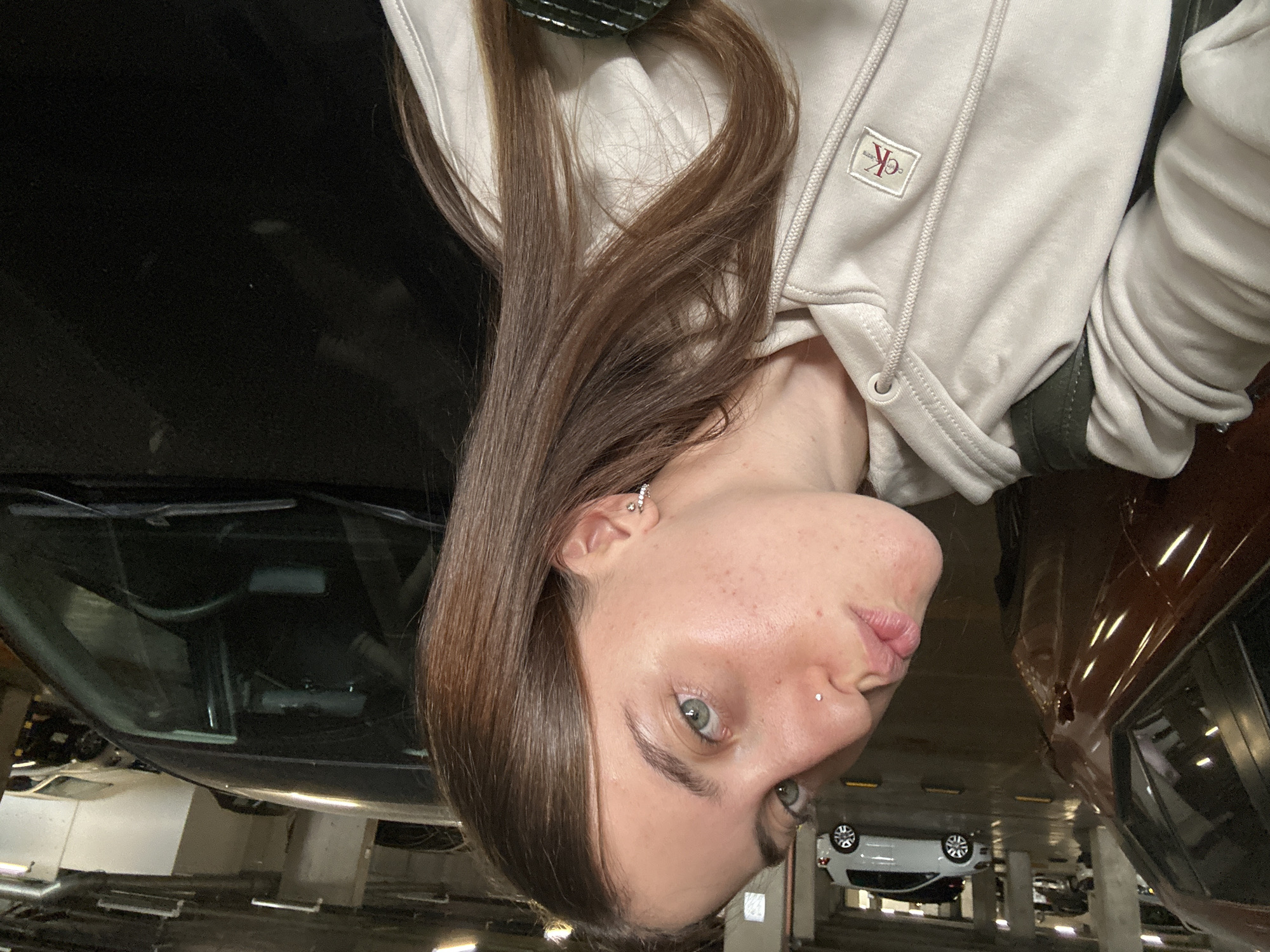 A selfie taken in an underground parking garage in Huixquilucan de Degollado, Mexico. A young adult female with light skin, light brown hair, and green eyes is the primary subject. She is wearing a light-colored hoodie with a "CK" logo. She has a nose piercing and earrings. Above and to the left of the subject is the underside of a dark-colored car, including a windshield and wiper blades. To the right of the subject is the side of a reddish-brown vehicle. Several white vehicles are in the background, parked between concrete support columns. The floor is concrete and shows evidence of staining. The lighting is provided by overhead fixtures, and there are other cars visible in the background. The image is rotated 180 degrees.