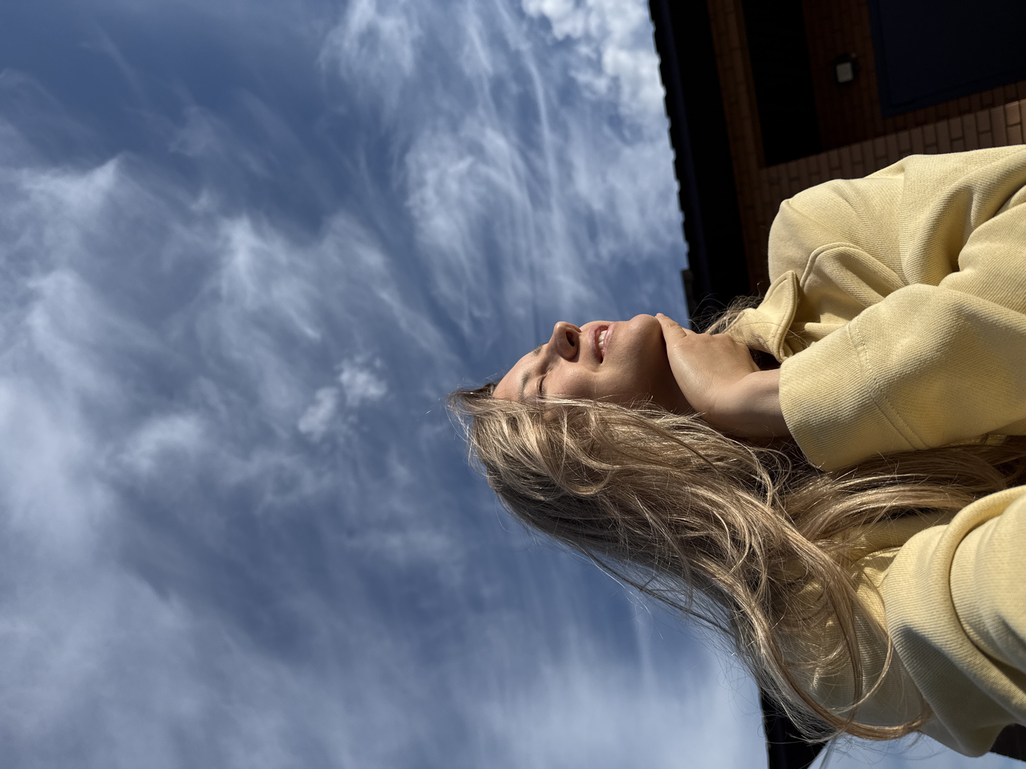 The image shows a young woman looking up at a blue sky with wispy white clouds. She has blonde hair and is wearing a yellow jacket. Part of a brick building is visible in the upper right corner. Based on the prompt, the location is Chaikovsky, Russia.