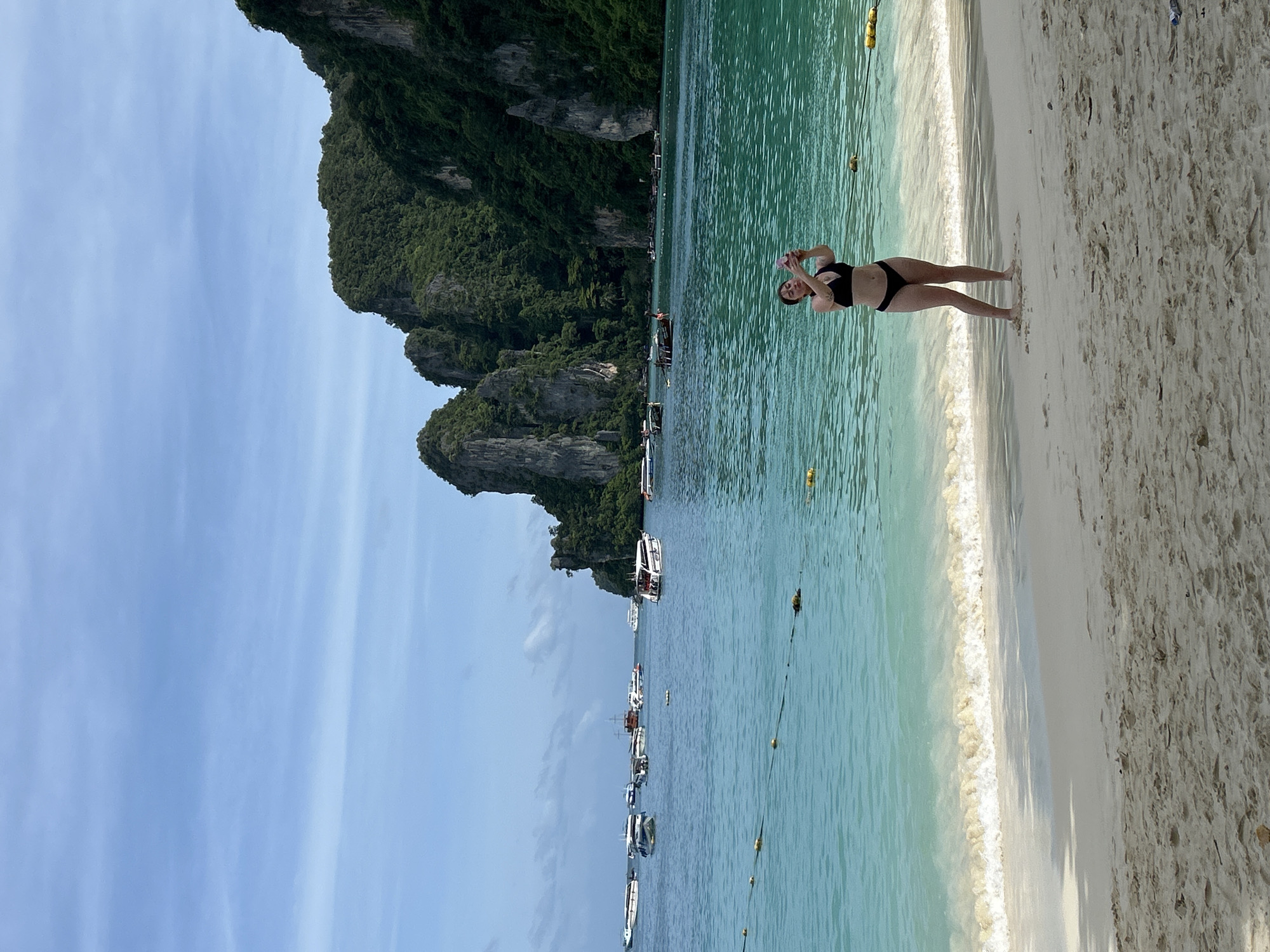 A woman in a black bikini stands on a sandy beach in Ao Nang, Thailand, holding a pink smartphone and taking a selfie. She is facing the ocean, which is a vibrant turquoise color. Behind her, several longtail boats and speedboats are anchored in the bay, and dramatic, lush green limestone karsts rise from the water. The sky is a pale blue with thin, wispy clouds. Gentle waves are breaking on the shore. The sand is light brown and appears wet near the water's edge. Yellow buoys are visible in the water, marking a boundary.