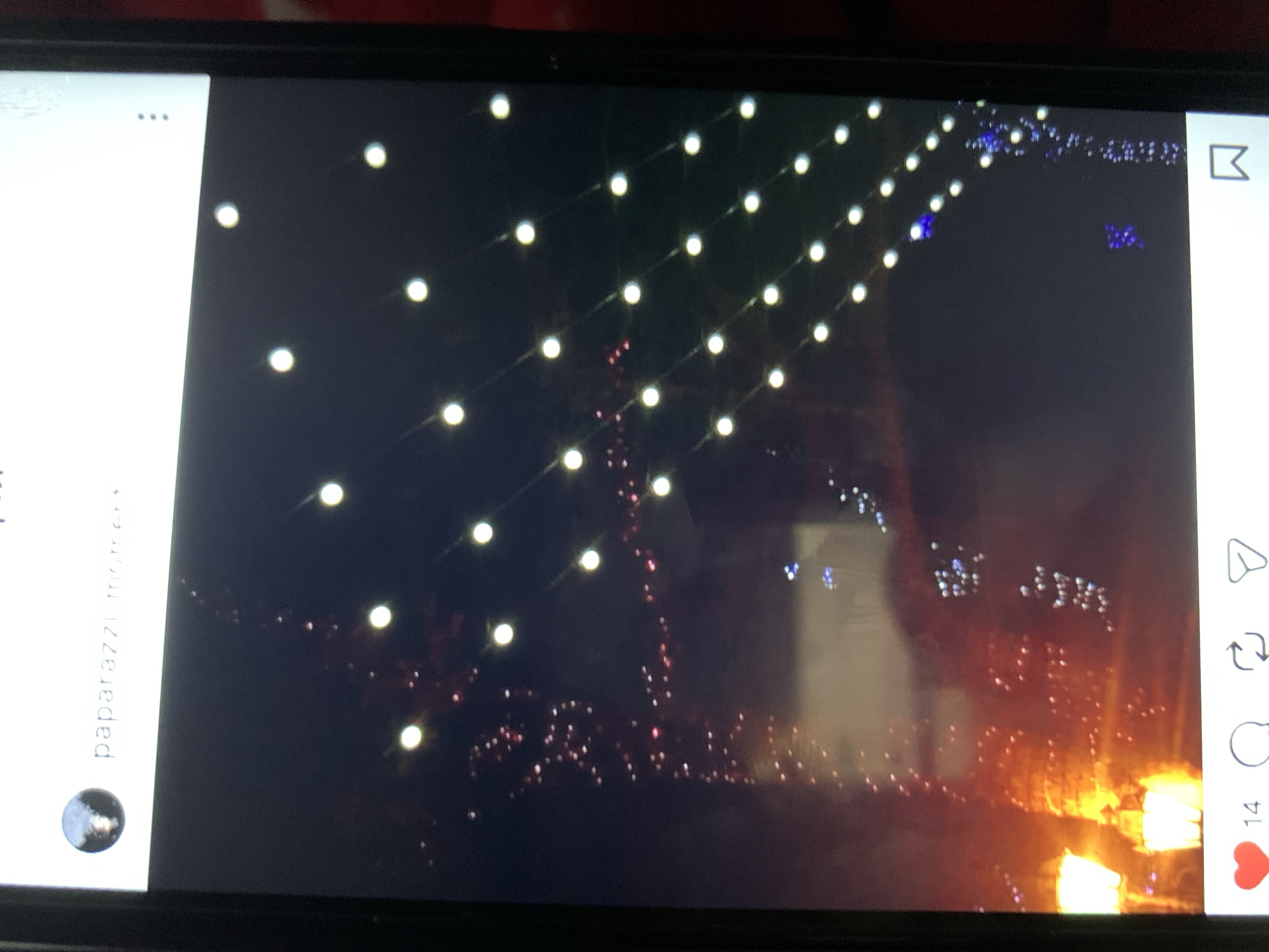 The image displays a close-up of a smartphone screen, exhibiting an Instagram post. The post features a dark, nighttime scene illuminated by numerous small, bright white lights arranged in diagonal rows, likely string lights. In the background, there are blurred hints of warm orange and red lights, possibly from streetlights or illuminated buildings, and some scattered smaller, colored lights. At the top left of the post, there is a profile picture and the text "paparazzi moment". At the bottom of the screen, social media interaction icons are visible: a red heart with the number "14" next to it, indicating likes, a speech bubble icon for comments, a refresh-like icon for sharing, and a bookmark icon. The image is taken from an angle, suggesting it's a photograph of a phone screen. No specific location in Lymanka, Ukraine, can be identified from the visual content of the post itself.