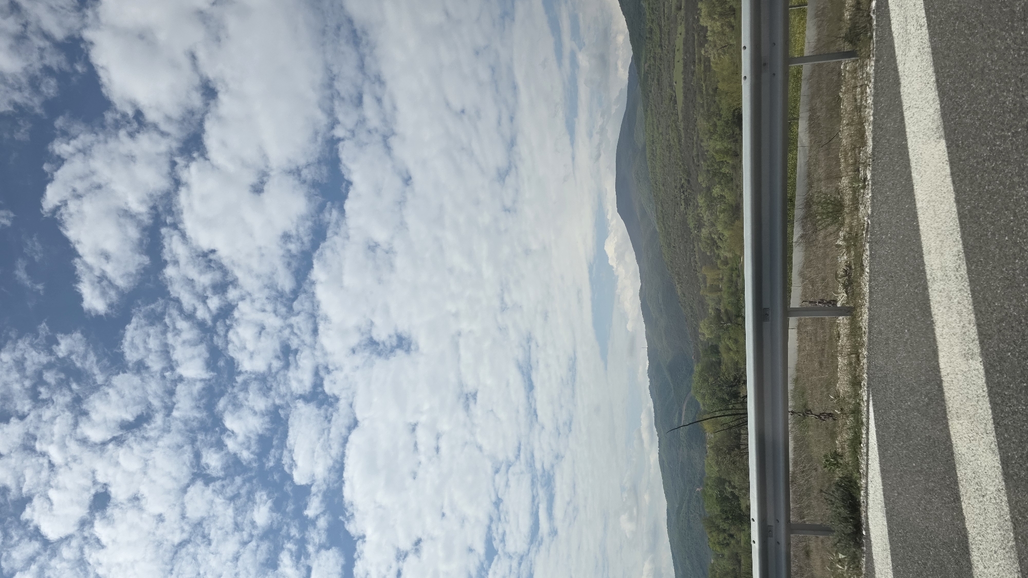 The image depicts a roadside scene with a view of mountains and a cloudy sky, likely taken on the outskirts of Valkovo, Bulgaria. The focus is on the edge of an asphalt road with a painted white lane marking and a metal guardrail. Beyond the guardrail, there is a grassy verge and a landscape of wooded hills, with mountains visible in the distance. The sky is predominantly blue, filled with numerous white clouds. There are no people visible.