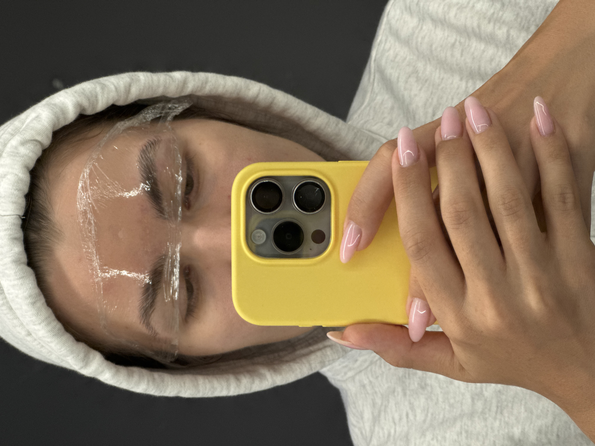A person wearing a light gray hoodie is holding a yellow phone up to their face, taking a selfie. The person's eyebrows appear to have a clear plastic film applied. Their nails are manicured with a light pink polish. The background is a dark gray, and the image appears to have been taken in Unknown, Unknown.