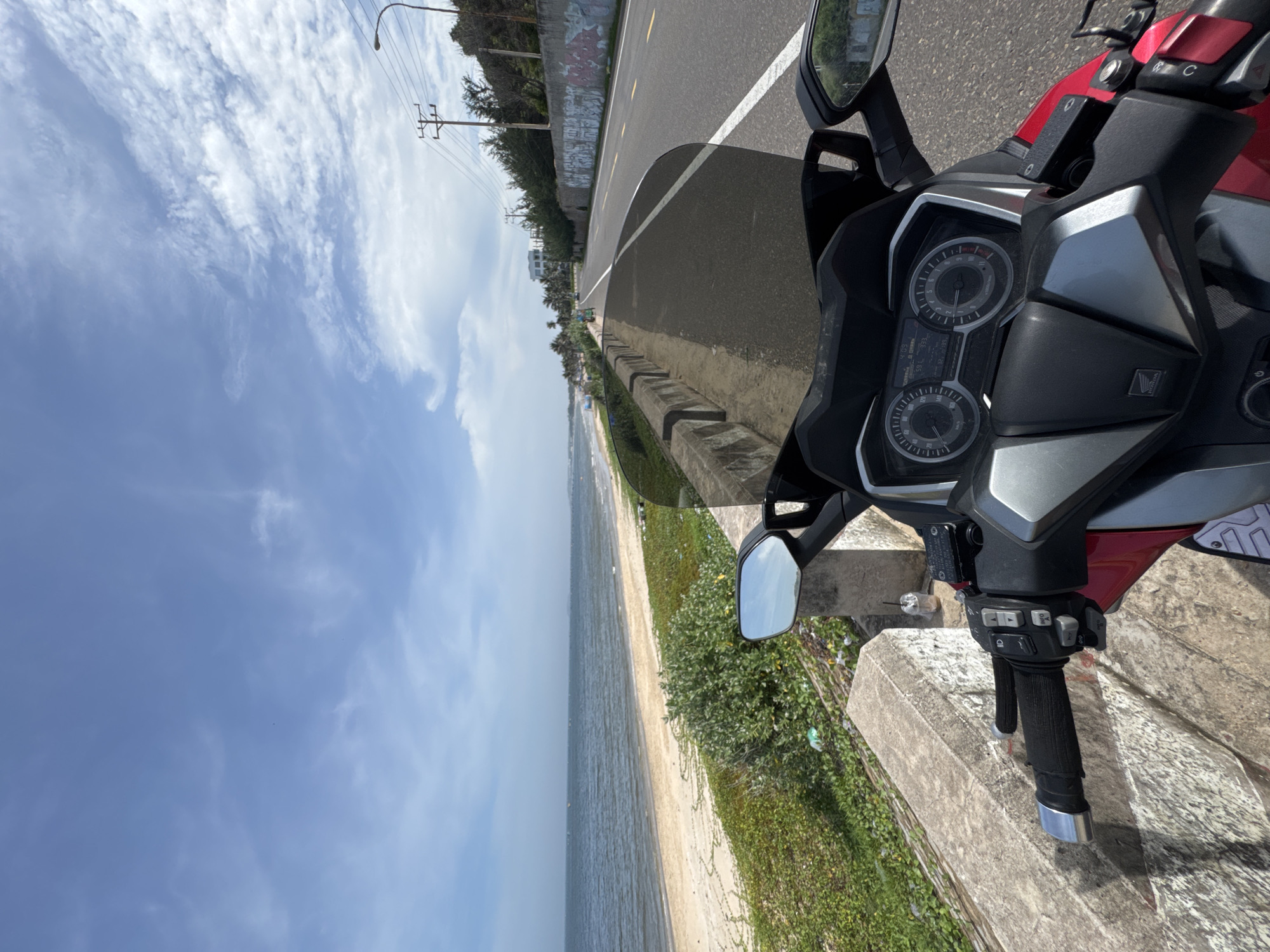 The image captures a view from the handlebars of a red scooter parked alongside a coastal road in Mui Ne, Vietnam. The scooter's dashboard, showing a speed of 59 km/h, is visible in the foreground. The road, marked with a white line, stretches ahead, bordered by a low concrete wall covered in graffiti on the right. To the left, a sandy beach meets the calm, blue ocean. Lush green vegetation grows along the edge of the road, with scattered litter visible among the plants. In the distance, the coastline is dotted with buildings and palm trees. The sky is a clear blue with scattered white clouds. A utility pole with electrical wires and a street lamp are visible on the right side of the road.