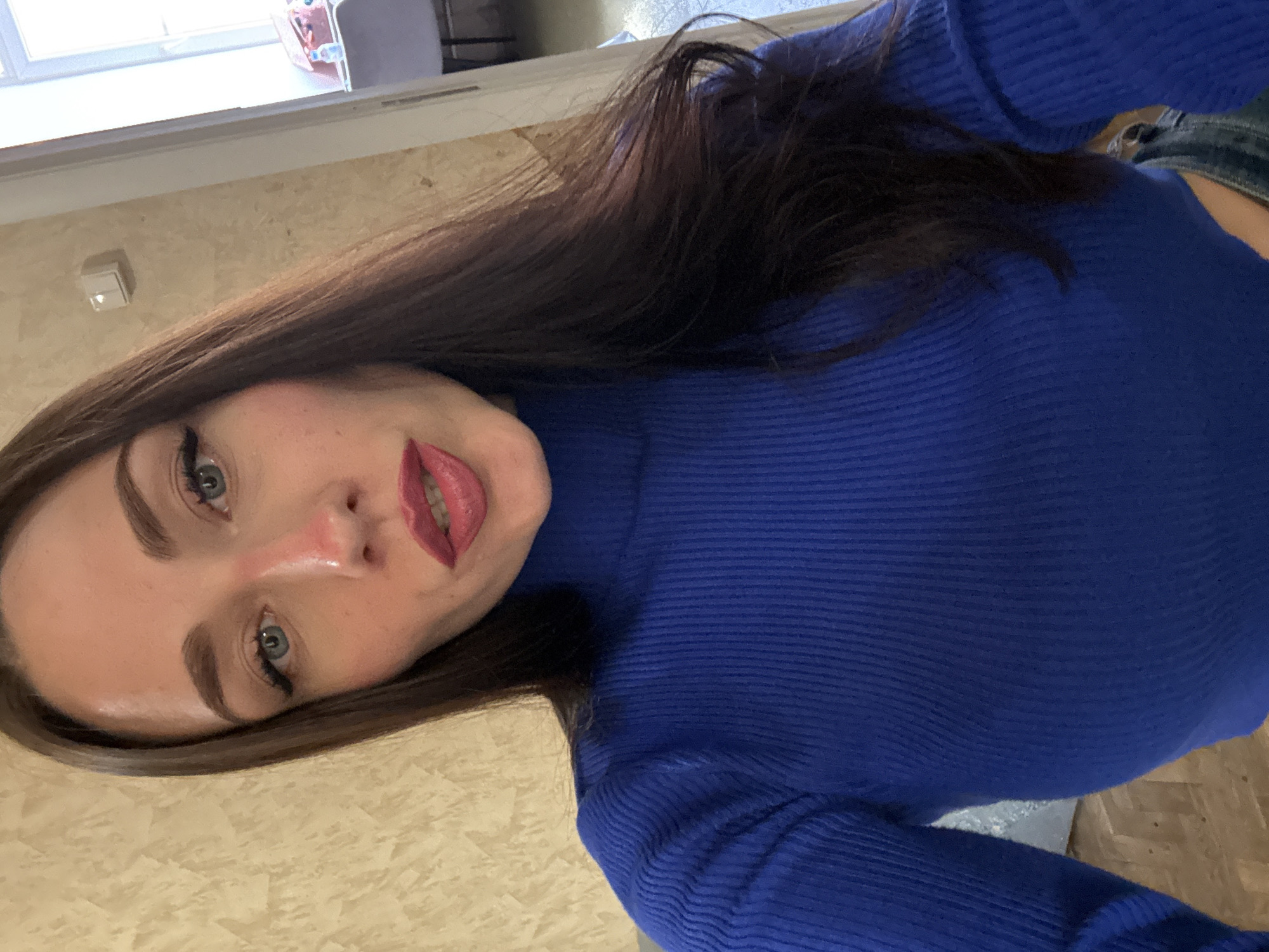 A young woman with long, dark hair and fair skin poses for a close-up selfie, her face angled toward the viewer. She wears a vibrant blue turtleneck sweater. Her makeup includes dark eyeliner and lipstick. The background shows a wall with an off-white patterned surface. The location is Saint Petersburg, Russia. A small, light-colored object is visible on the wall near the top left. A glimpse of jeans is visible.