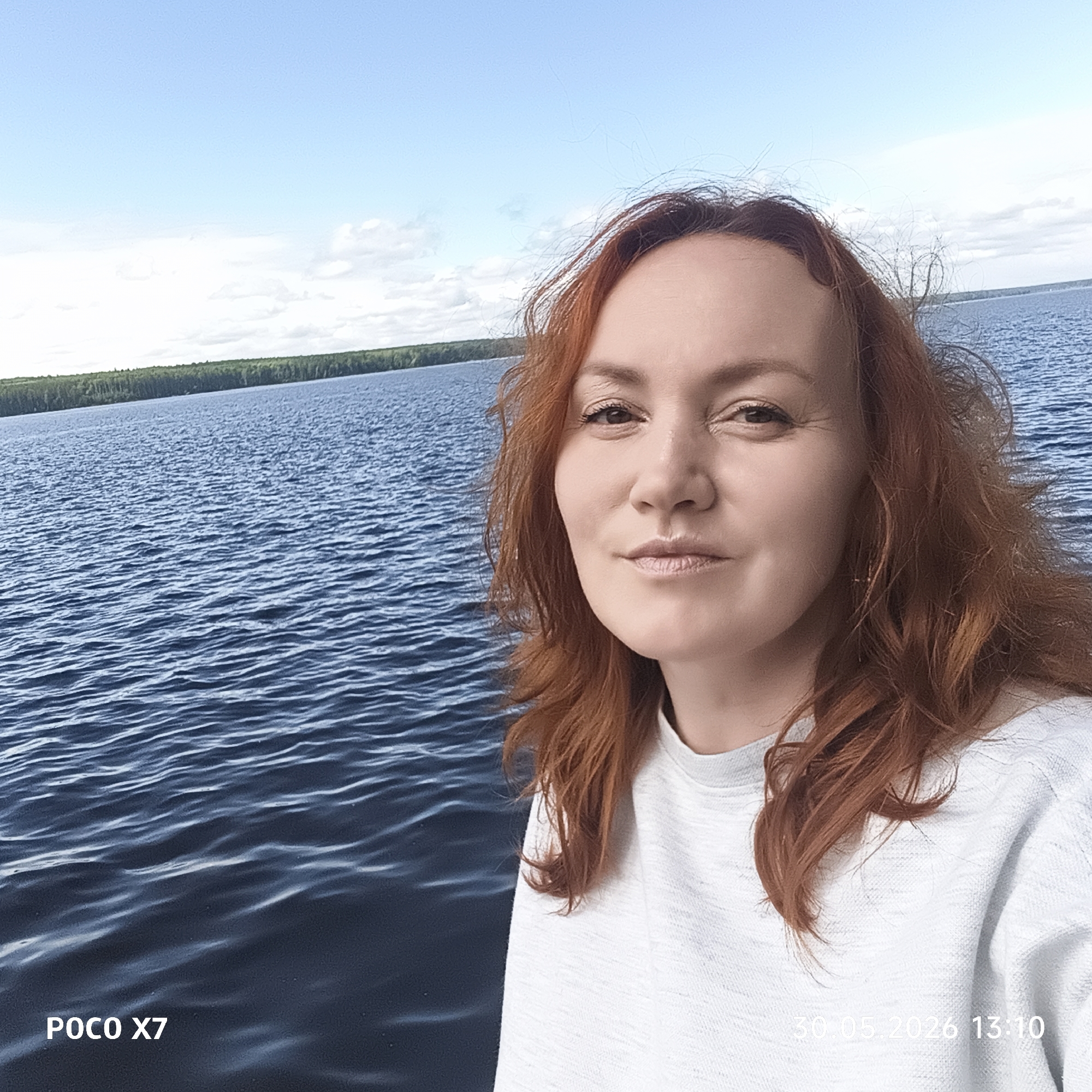 A selfie captures a woman with red hair, possibly standing near a body of water, likely the Kama River in Chaikovsky, Russia. The water appears rippled, extending to a visible shoreline with trees in the distance. The sky is partly cloudy. The woman is wearing a white sweater and is positioned on the right side of the frame. Text at the bottom of the image displays "POCO X7" and "30.05.2026 13:10," indicating the camera and date/time.