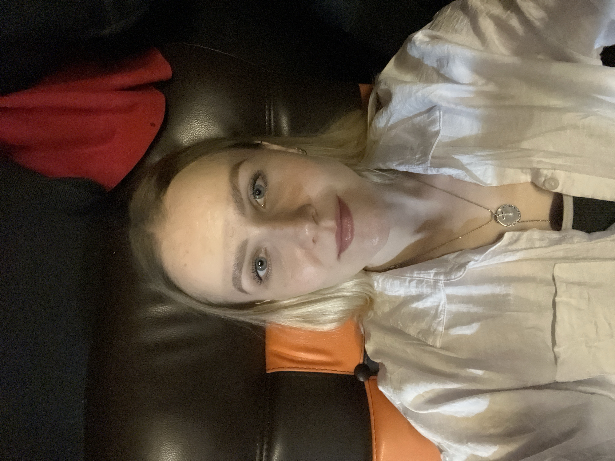 A selfie image shows a woman with light skin, blonde hair, and blue eyes. She's wearing a white button-down shirt and a silver necklace with a round pendant. She's lying on a dark brown and orange leather chair or sofa, with a red cloth visible in the background to the left. The location is not evident from the image, and there is no indication of Khimki, Russia.