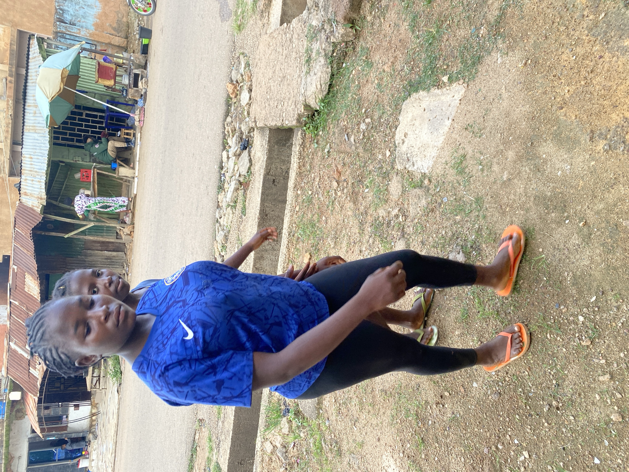 Two young girls are the main subjects in this vertically oriented image, set in an outdoor, urban environment in Jos, Nigeria. The girl in the foreground, facing slightly away from the camera, wears a bright blue t-shirt with a white Nike logo and a subtle pattern, paired with black leggings. Her hair is styled in braids. Behind her and slightly to her left, another girl is visible, also looking towards the camera.
They are standing on a dirt and gravel ground, with patches of sparse green grass. To their left, a portion of a corrugated metal structure, possibly a makeshift shop or dwelling, is visible. An umbrella, partially open and colored in light blue and tan, is propped up near this structure, suggesting it might be providing shade. A paved road runs alongside the scene.
The overall impression is that of a casual, everyday moment. The lighting suggests it is daytime, likely not harsh midday sun given the shadows. The girls appear calm and perhaps curious, with the foreground girl's gaze directed downwards and slightly to the side. There are no clear indications of specific events or strong emotions. The background shows a mixture of weathered buildings and the natural elements of the roadside, typical of many urban areas in Nigeria.