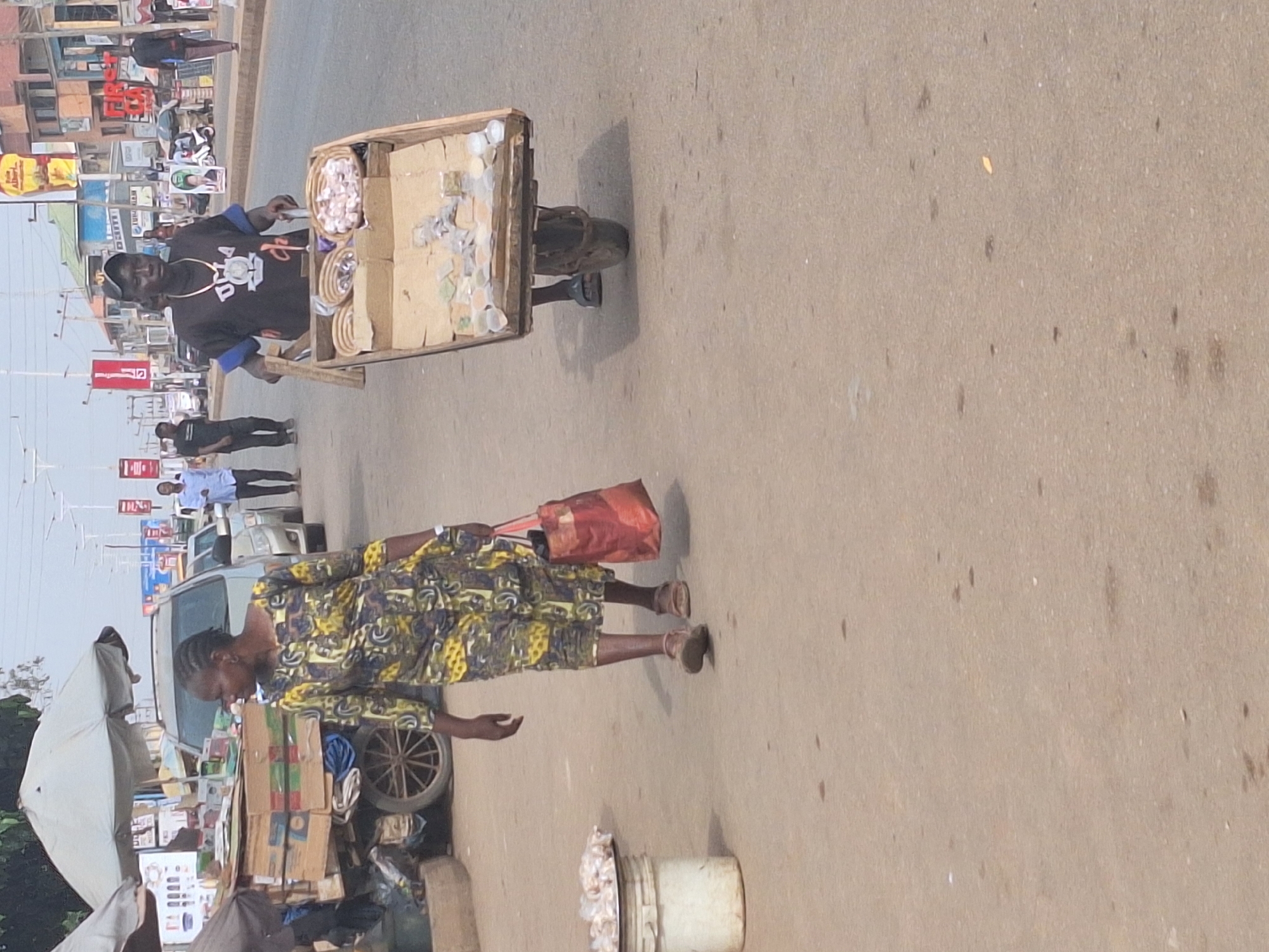 The image depicts a street scene in Sabon Tasha, Nigeria. A man wearing a black t-shirt and baseball cap is pulling a cart loaded with goods. A woman in a patterned dress walks nearby, carrying a red handbag. Other people and a vehicle are visible in the background, along with signs and merchandise indicating a commercial area. A bucket of goods sits on the ground near the curb.