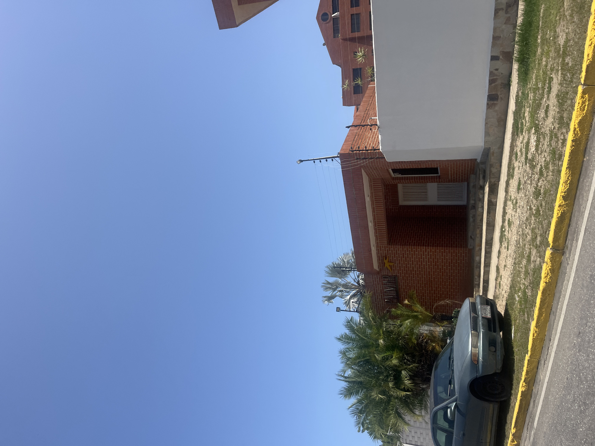 The image shows a clear blue sky over Pampatar, Venezuela, on a sunny day. In the foreground, a dark gray sedan is parked on the side of a road with a yellow curb. The car faces to the right and has a license plate that reads "AAS5HGO". Behind the car is a building constructed of red brick. The building has a covered entryway with stairs leading up to a white door with a metal grate. Above the door, the word "PALMAR" is visible in raised letters, along with a yellow stylized palm tree logo. Palm trees are visible to the left of the building. In the background, a multi-story brick building with dark windows can be seen. The scene appears to be a street in a residential or commercial area.