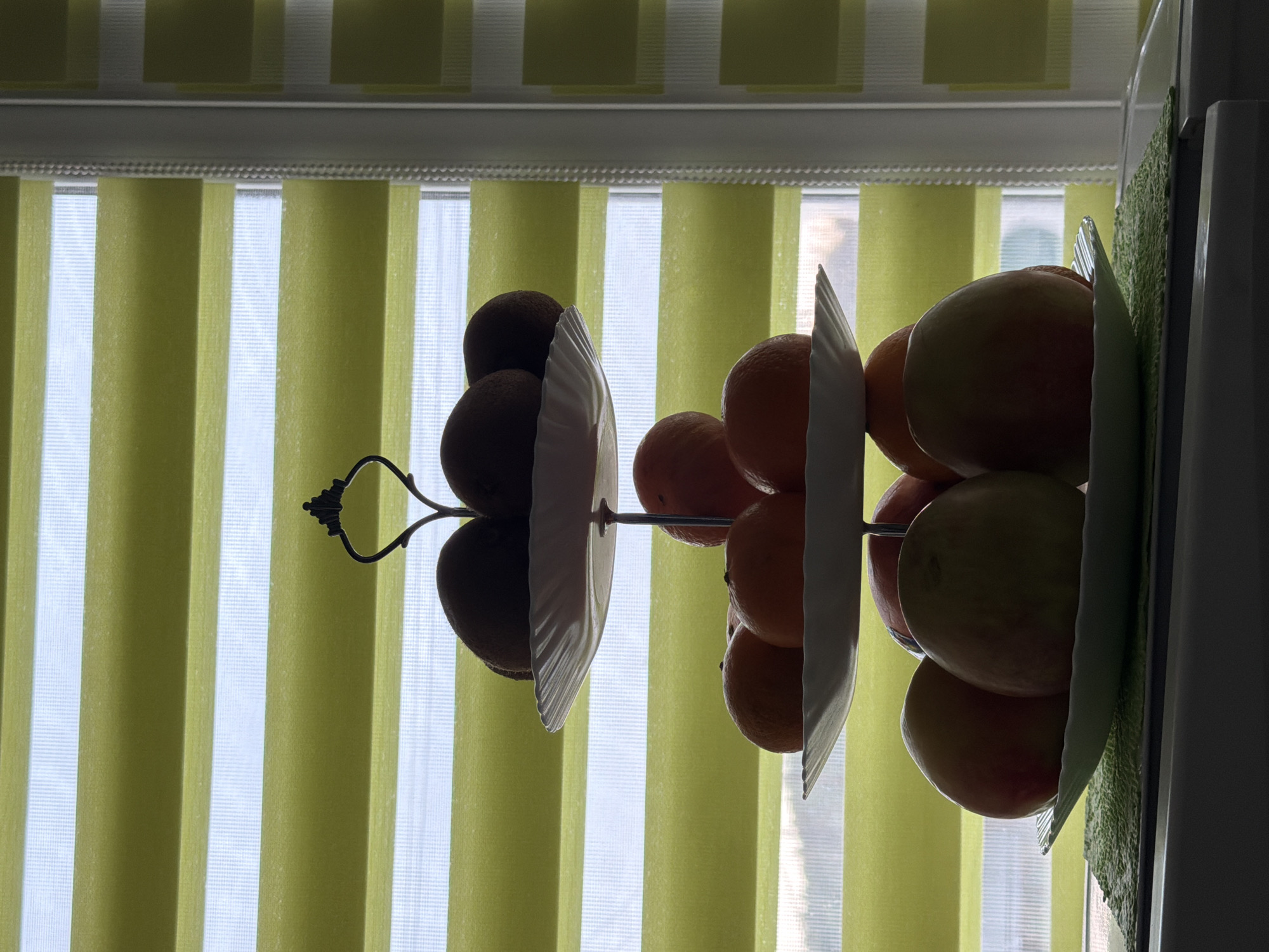 A two-tiered fruit stand is positioned in front of a window with vertical blinds. The fruit stand holds kiwis on the top tier, oranges in the middle, and apples on the bottom. The blinds are mostly closed, creating alternating stripes of light and shadow on the wall behind the fruit stand. The background suggests an interior setting, possibly in Chaikovsky, Russia, due to the information provided, though there is no direct evidence in the image itself. The scene appears still, with no visible action or interaction taking place.