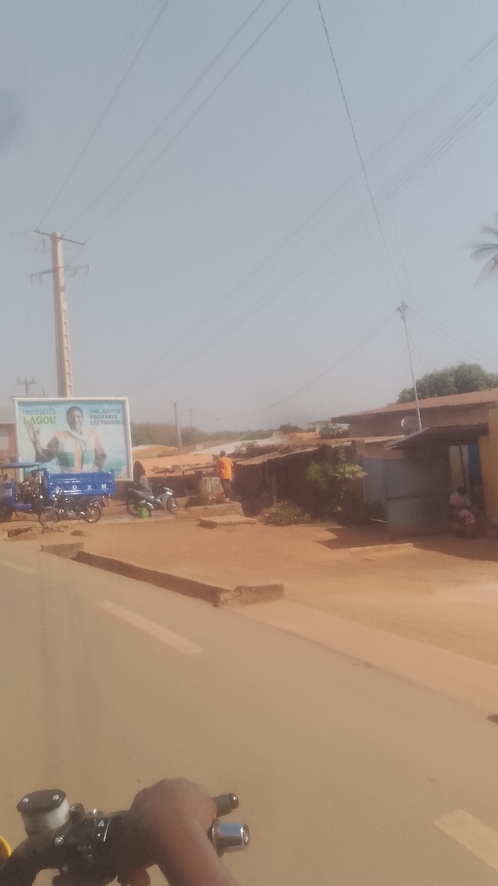 The image depicts a daytime street scene in Korhogo, Côte d'Ivoire, observed from the perspective of a motorcycle rider. A dark-skinned hand grips black handlebars, featuring a visible brake fluid reservoir and chrome throttle end, in the lower-left foreground. The sky is hazy and light blue.
A paved road with faded white lane markings extends into the midground, flanked by reddish-brown dirt and raised concrete curbs. On the left, a tall utility pole supports multiple overhead electrical wires that traverse the frame. Adjacent to the pole stands a large billboard displaying a smiling woman identified as "Henriette LAGOU," with the text "UNE JUSTICE EQUITABLE EST POSSIBLE." Parked beneath this billboard are a blue three-wheeled cargo vehicle, marked "DXM," and two motorcycles, one blue and white, the other silver and black.
On the right side of the road, a series of single-story structures with corrugated metal roofs line the street, appearing to be commercial or residential buildings. One individual, wearing an orange shirt, stands outside one of these structures in the midground. Further right and recessed within an open-fronted structure, two additional individuals are visible. Sparse vegetation and additional utility lines are present in the background and above the structures. A palm frond is partially visible on the far right against the sky.
