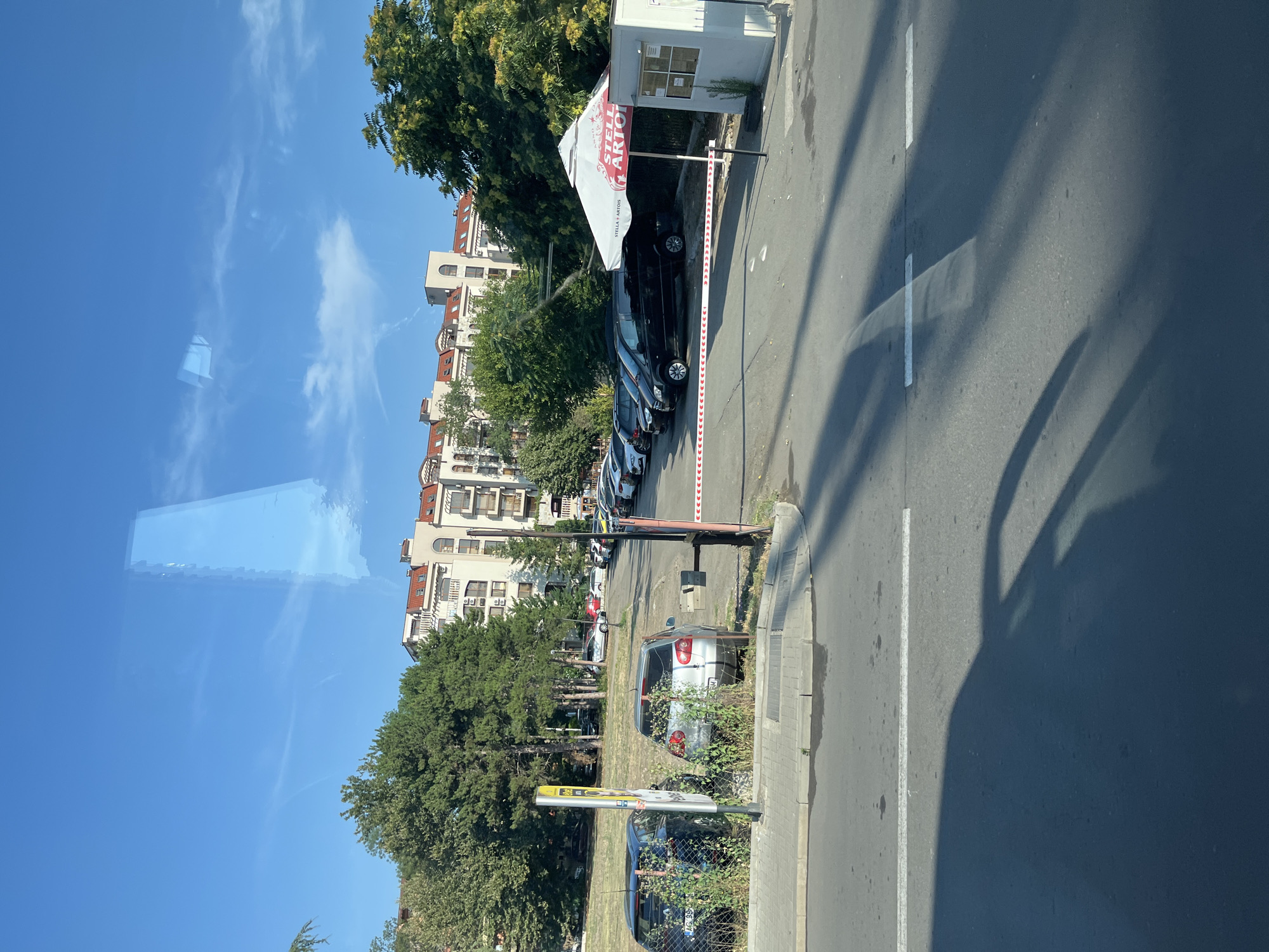 The image depicts a parking area with a barrier system and a security booth, located in the city of Nesebar, Bulgaria. In the foreground, a paved road is visible with tire marks and shadows. Beyond the road, a sidewalk and a parking lot are seen. Several cars are parked in the lot, including a dark-colored sedan and a silver hatchback in the foreground. Further back, a row of black vans and cars are parked, extending towards a security booth. The security booth is a small, white structure with a window and a red and white striped barrier arm extended across the entrance. A large white umbrella with the "Stella Artois" logo is set up next to the booth. In the background, a multi-story building with terracotta tiled roofs and balconies is visible, surrounded by green trees. The sky is blue with some wispy clouds. The overall scene suggests a secure, managed parking area in an urban or resort setting.