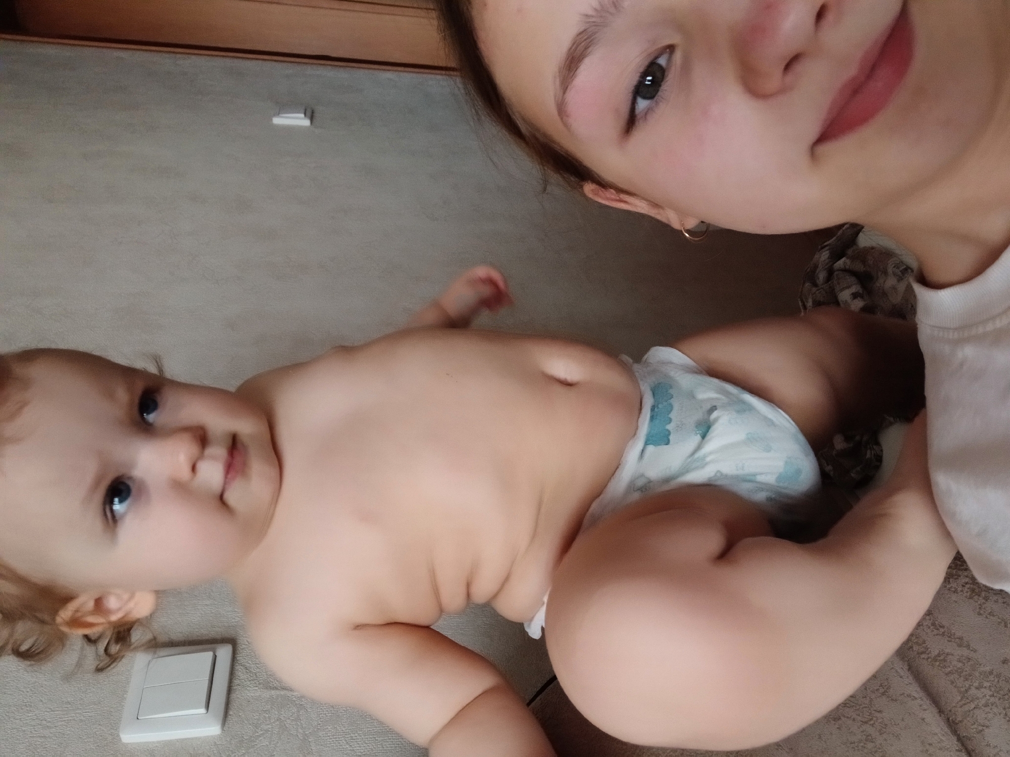 A photo taken in Krasnodar, Russia, presents two individuals: an adolescent girl and a baby. The adolescent girl is in the upper right quadrant of the frame. The baby, wearing a diaper, lies near a light switch in the bottom left portion of the image. The background appears to be a bed or a padded surface.