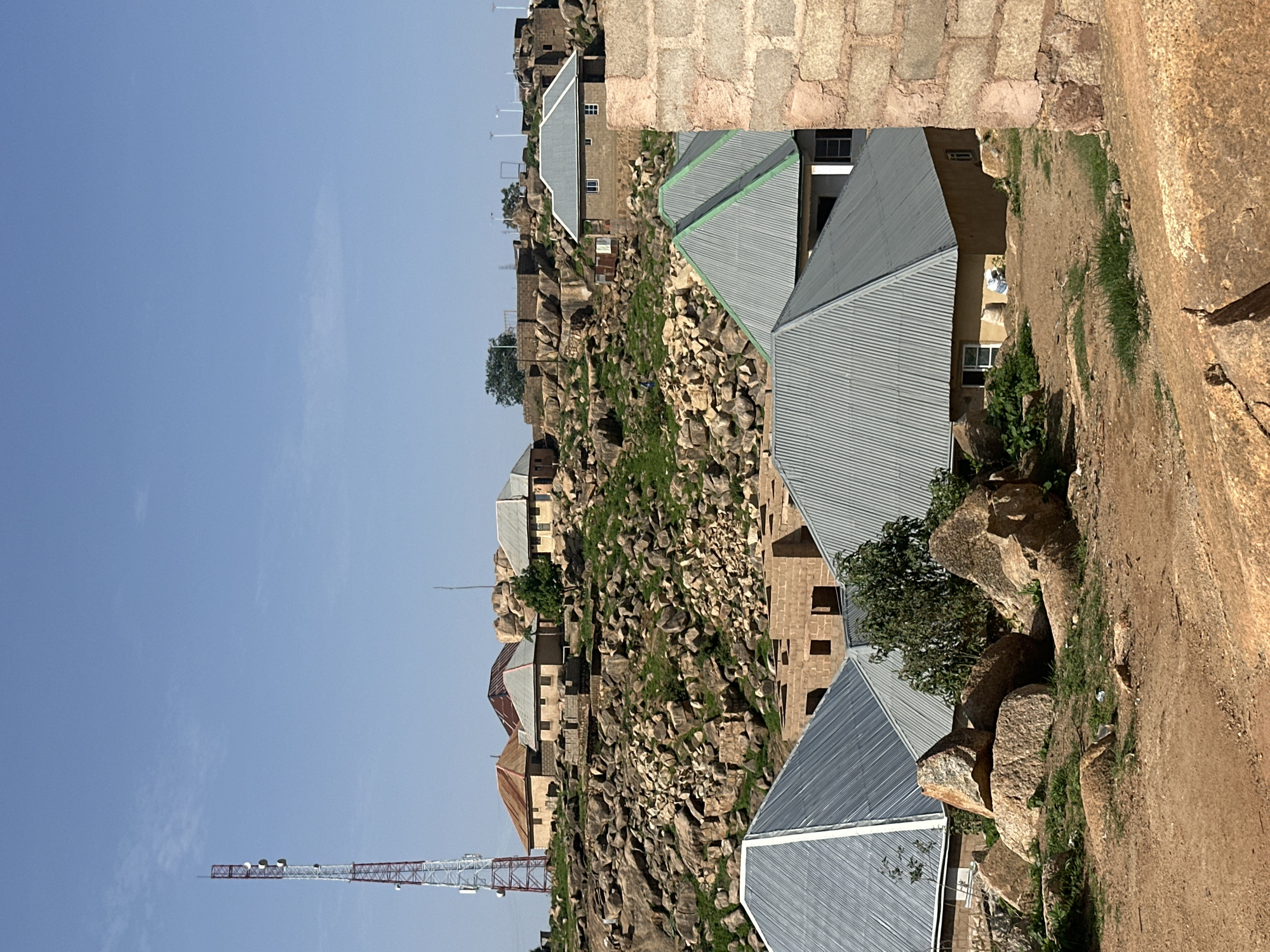 The image depicts a scene in Jos, Nigeria, showcasing a settlement nestled among rocky terrain. A prominent red and white telecommunications tower stands tall against a clear blue sky on the left side of the frame. Scattered across the rocky landscape are several buildings, characterized by their light-colored walls and predominantly corrugated metal roofs, some with green accents. Some structures appear to be under construction, with exposed brickwork. The ground is a mix of dirt, rocks, and sparse green vegetation. In the foreground, large boulders are visible, along with some scattered debris. The overall impression is of a functional, if somewhat rugged, residential area situated in a naturally rocky environment. No people are clearly discernible in the image, though the presence of buildings suggests habitation.
