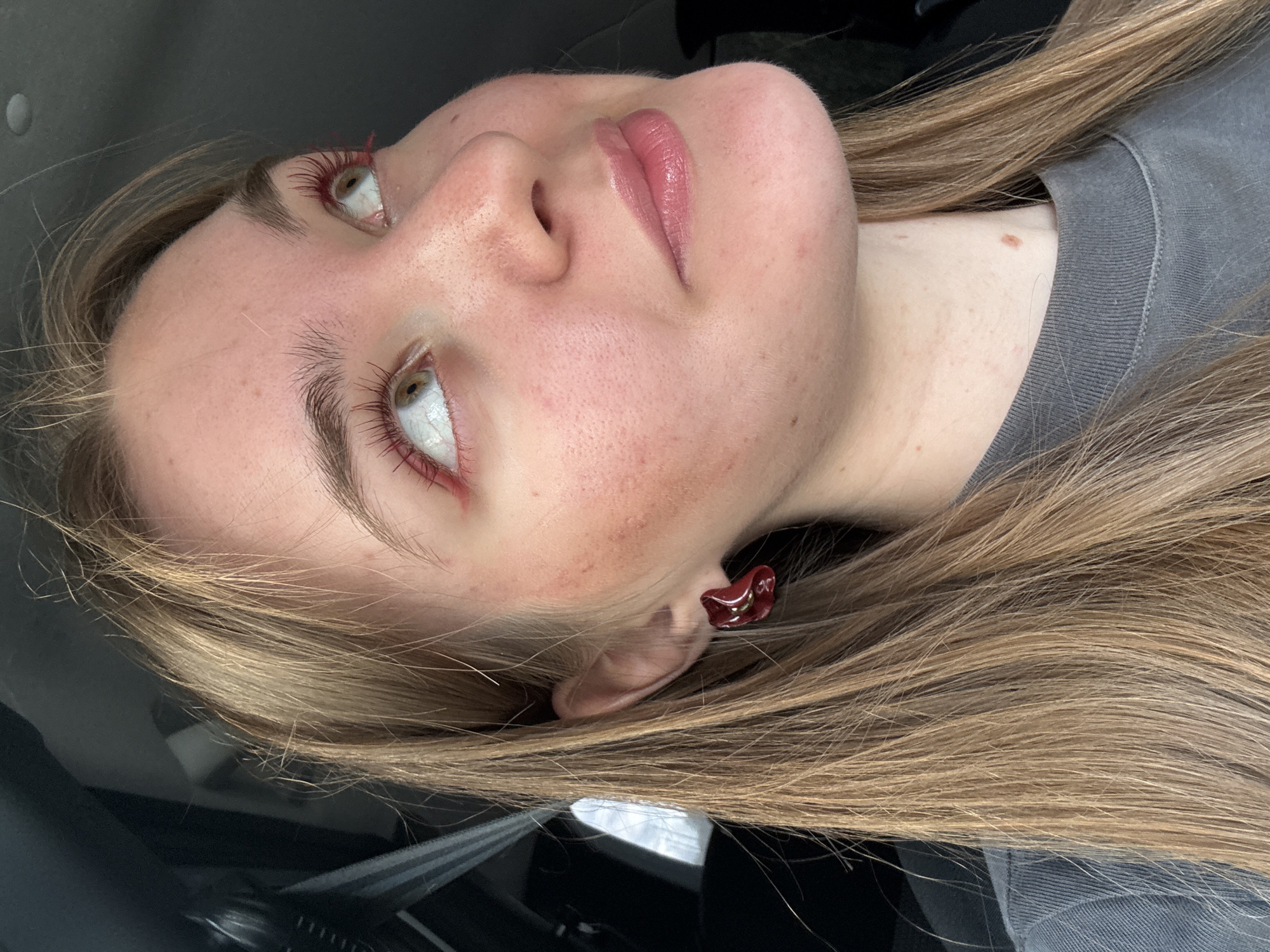 A medium shot of a young woman with long blonde hair is shown from the chest up. She is looking up and to the right, with her head tilted slightly. She is wearing a dark gray t-shirt and a dark red earring on her left ear. Her eyes are light brown, and she is wearing red mascara and a subtle pink lip gloss. Her skin is fair with some redness and freckles. The background is dark and out of focus, suggesting she is inside a vehicle or a dimly lit indoor space. The image does not contain information about the city of Perm, Russia.