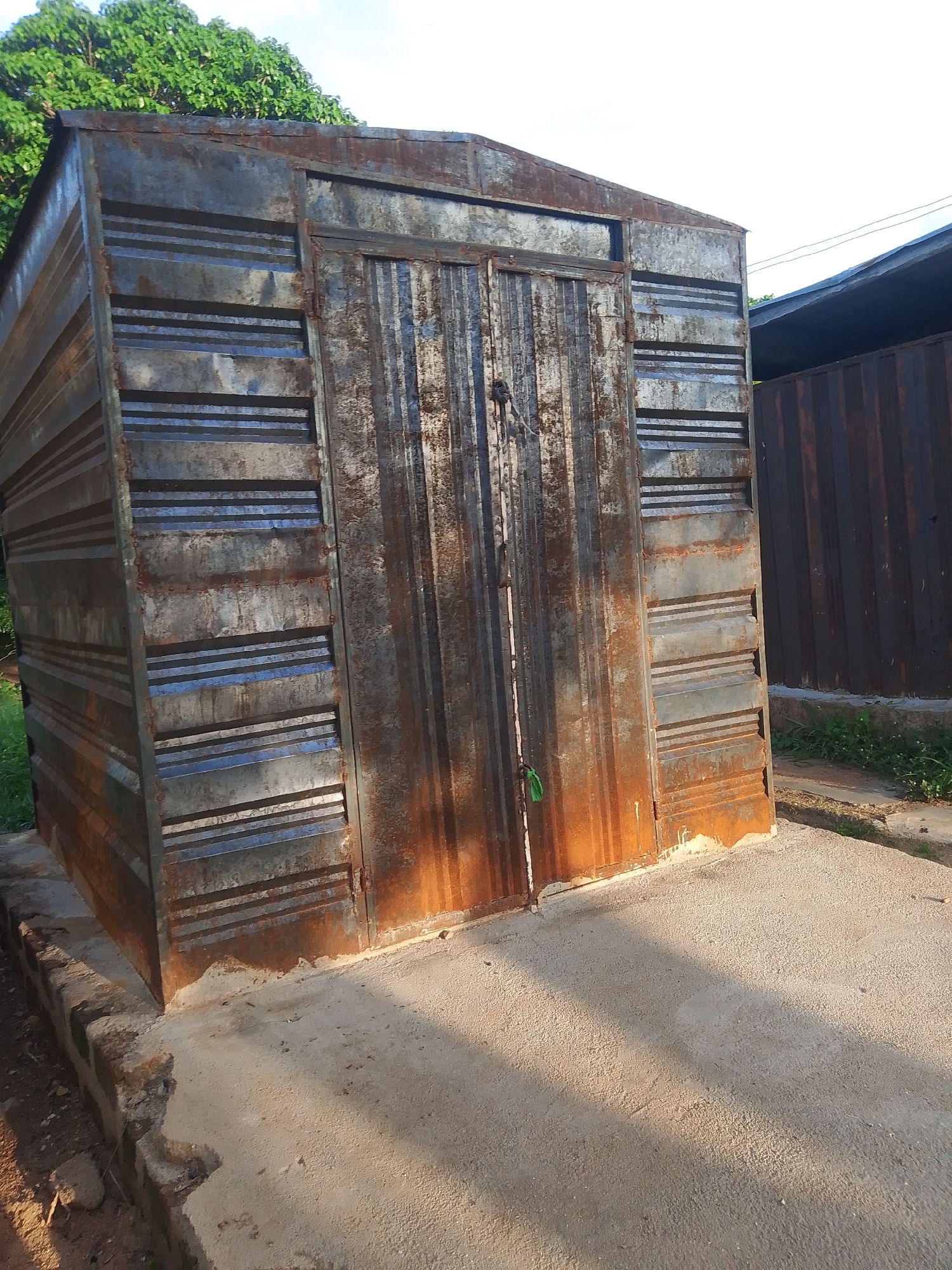 This is a medium-shot, eye-level, outdoor photograph of a small, rectangular, corrugated metal structure with a double door. The metal of the structure is heavily rusted, showing a mix of metallic grey, orange, and brown hues. The door is also made of corrugated metal and appears to be secured with a simple latch and possibly a padlock. A small piece of green material is tied to one of the doors.
The structure is situated on a concrete slab, which is uneven and shows signs of wear. To the left of the structure, there's a low-lying brick wall, partially crumbled. Behind the structure and to the right, another corrugated metal fence or wall is visible, also showing signs of rust. Lush green trees are visible above and behind the metal structure on the left, suggesting a vegetated area. The ground around the concrete slab is a mix of dirt and sparse grass.
The lighting suggests it is daytime, with shadows cast on the concrete slab, indicating the sun is at an angle, possibly late afternoon. The sky is partially visible at the top of the image and appears to be a pale blue or white, with hints of clouds or haze. There are some power lines visible in the upper right portion of the frame. The overall impression is of a functional, albeit weathered, utility structure in an outdoor setting. No people are visible in the image. No text is discernible.