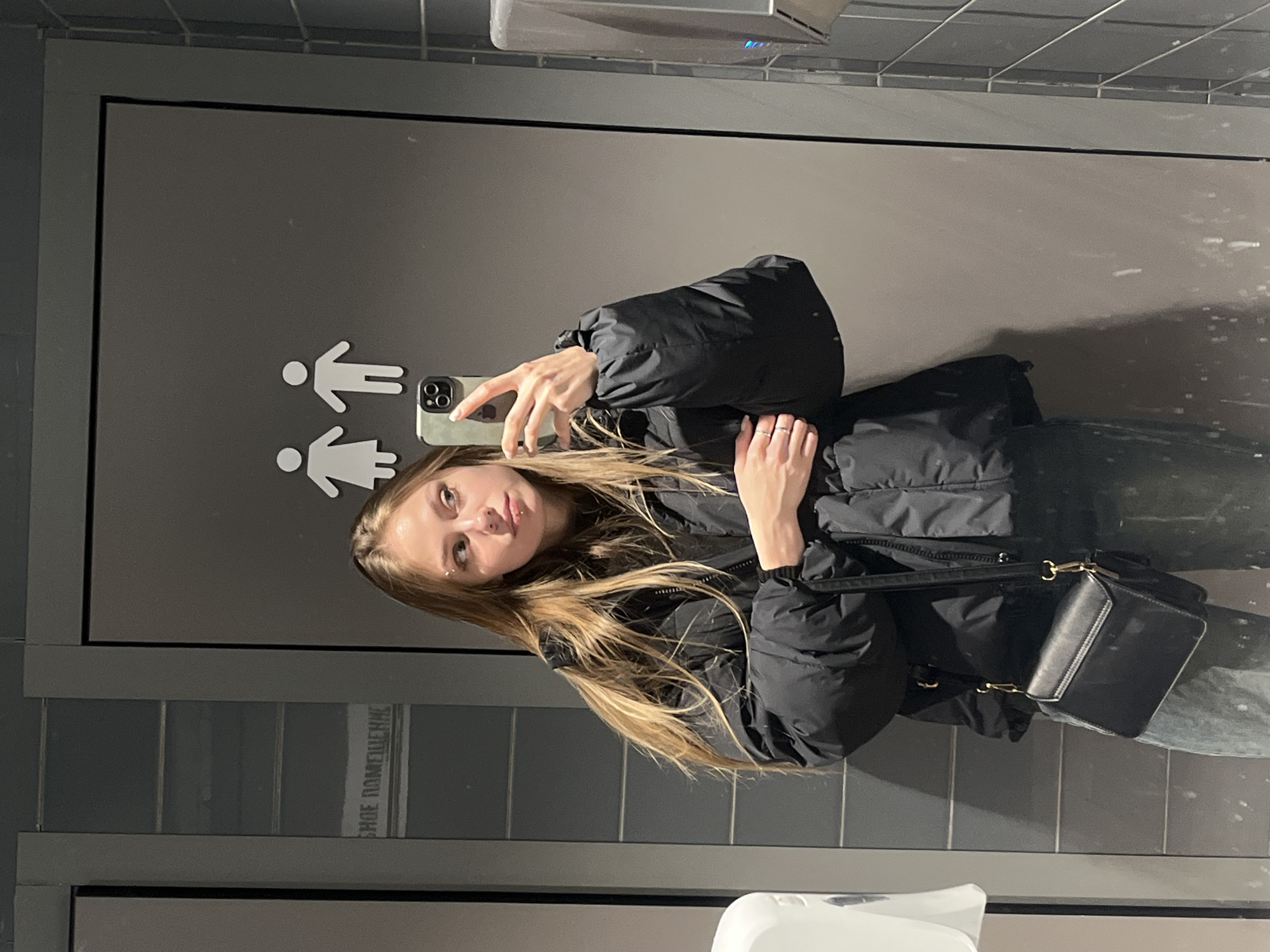 A blonde female takes a selfie in a bathroom in Chaikovsky, Russia. The mirror reflects bathroom signage and tiled walls. The female wears a black puffy jacket and carries a black purse.