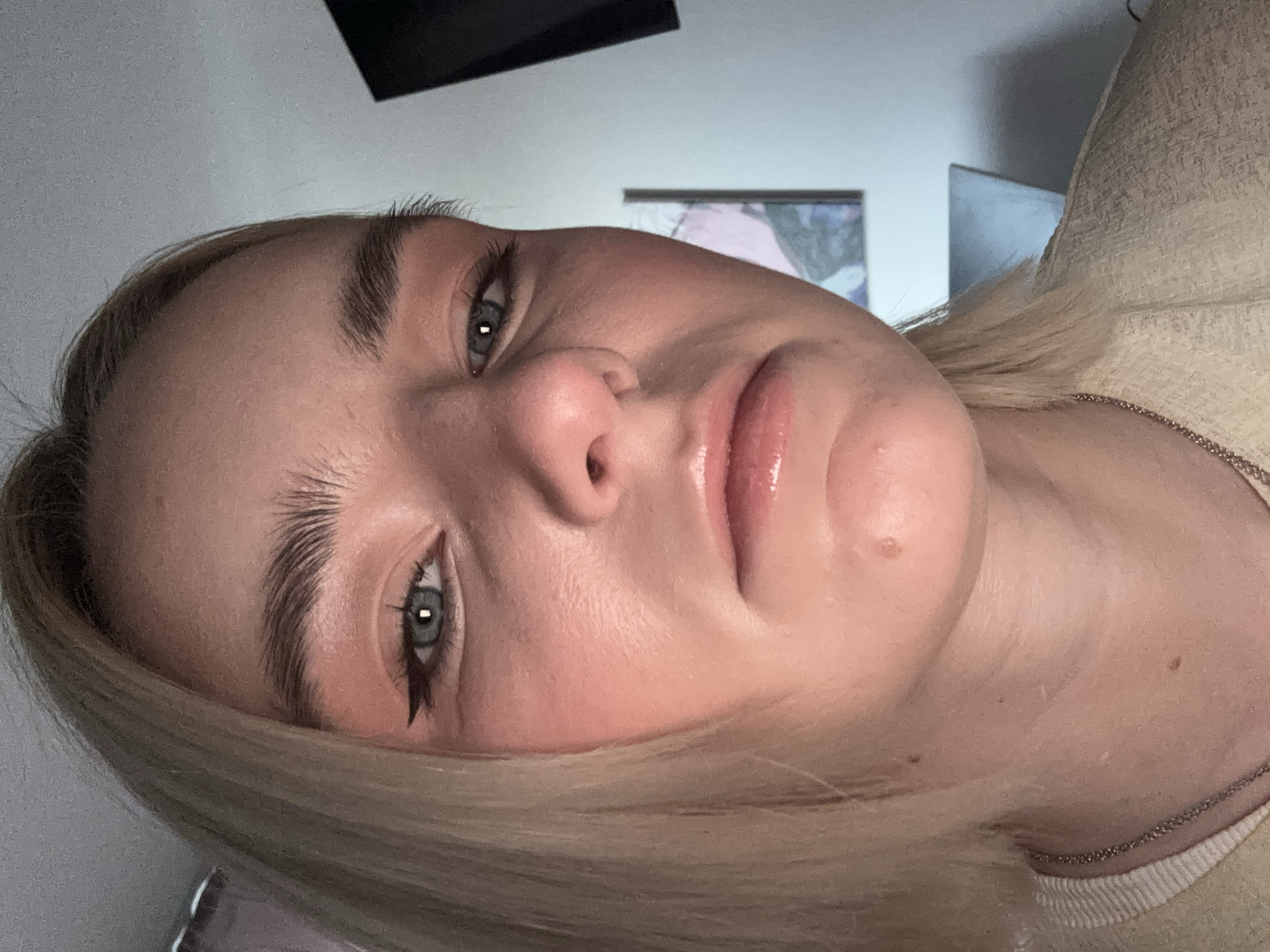 A close-up, tilted portrait of a light-skinned female with blonde hair. She has light blue eyes with dark eyeliner on the upper lids. Her eyebrows are thick and well-defined. Her lips are glossy. A small mole is visible on her lower right cheek. She is wearing a light-colored top. A thin silver chain necklace is visible around her neck. The background consists of a white wall with two dark, rectangular objects and a framed picture. The location of the image is unspecified.