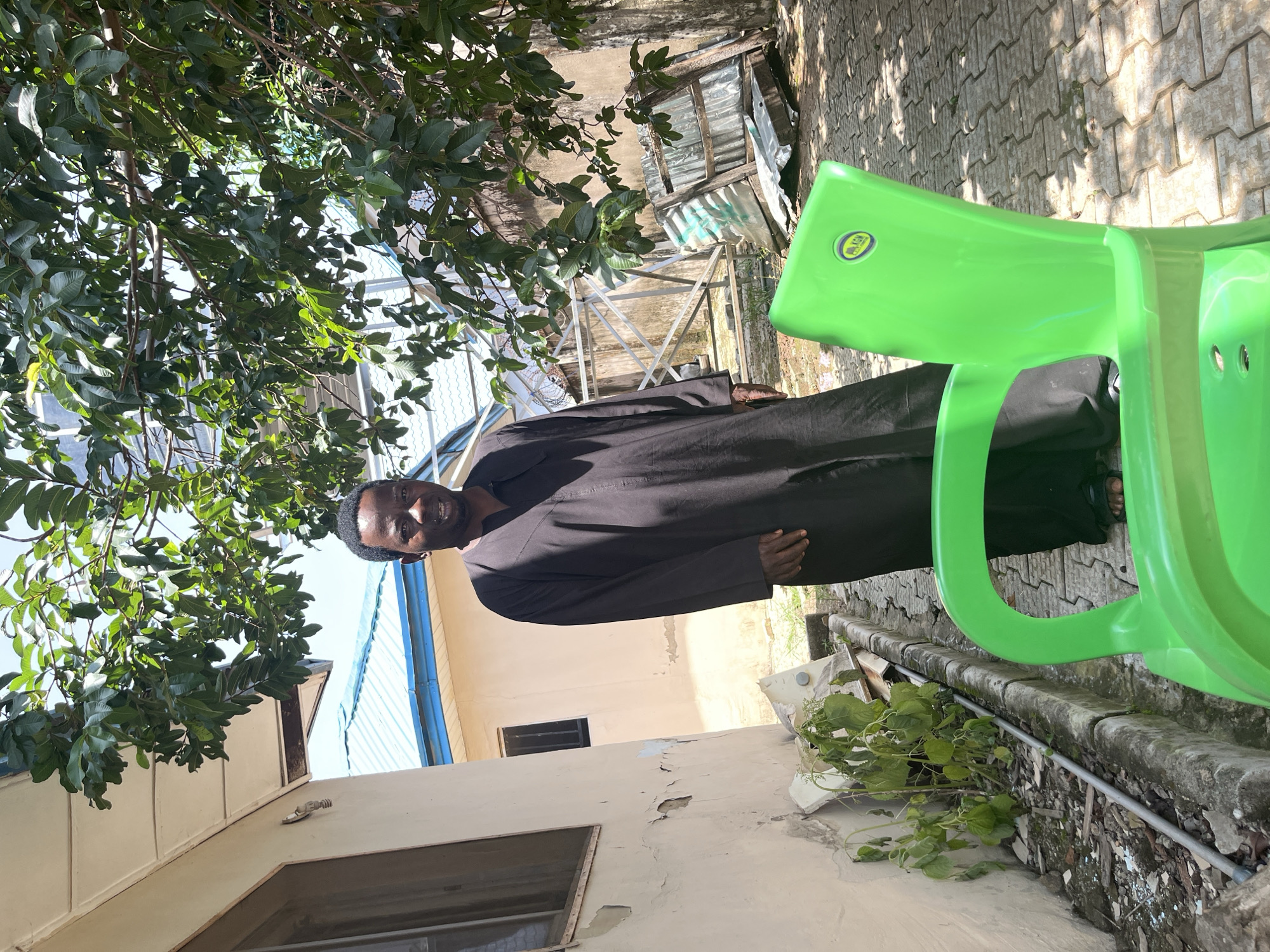 A Black man stands in what appears to be a residential courtyard in Abuja, Nigeria. He is wearing a long, black, traditional garment and is looking towards the camera. In the foreground, a bright green plastic chair is positioned prominently, partially obscuring the man's lower body. The man is standing on a paved surface made of interlocking concrete blocks. To the left of the man is a light-colored building with a textured, peeling facade and a window. To the right, there is a wooden structure with corrugated metal roofing leaning against a wall. Overhanging the scene are the green leaves and branches of a tree, casting dappled shadows on the ground and structures. The background reveals more buildings with blue roofing, suggesting an urban or suburban setting. The overall impression is a candid outdoor shot in a somewhat weathered, everyday environment.