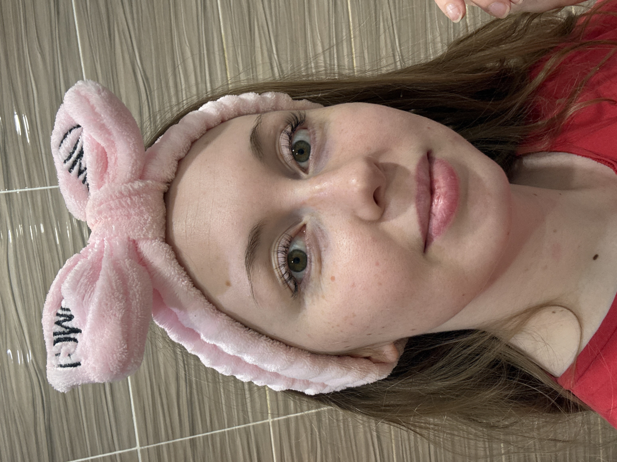 A young woman with long brown hair and greenish-hazel eyes looks directly at the viewer in this portrait, which is oriented with her head tilted to the right. She has a fair complexion with visible freckles and her expression is neutral to slightly contemplative. Her lips are full and appear to have a natural or light pink tint. Her eyelashes are long and well-defined.
She is wearing a soft, pink, fuzzy headband with a large bow on top, which helps keep her hair back from her face. The letters "OMG" are embroidered in black on the bow of the headband. She is dressed in a red top. In the background, there is a tiled wall with rectangular, light-colored (possibly grey or beige) tiles arranged vertically, suggesting an indoor setting, potentially a bathroom or a similar area for personal care. A hand with manicured nails is partially visible at the top of the frame, near her hair.