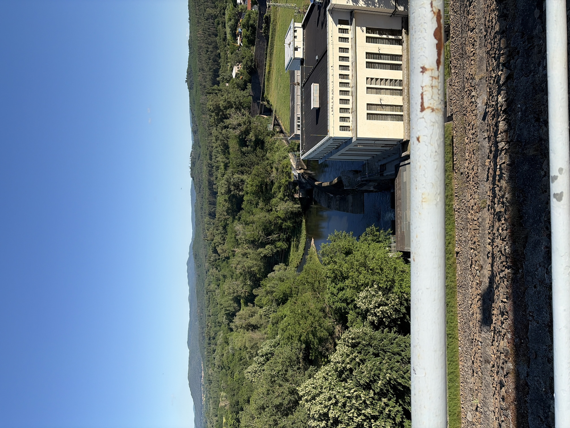 This is an eye-level, high-angle, medium shot of the Koprinka Dam in Koprinka, Bulgaria, taken on a clear, sunny day. The foreground is dominated by a white, weathered metal railing. Below the railing, a rocky embankment transitions into a concrete structure. The middle ground features a large, cream-colored dam building with a black roof, characterized by rows of tall, narrow windows. Adjacent to the building, a dam spillway releases water into a river that winds through a lush, green forest. The background showcases rolling, tree-covered hills under a bright blue sky. No people are visible in the frame.