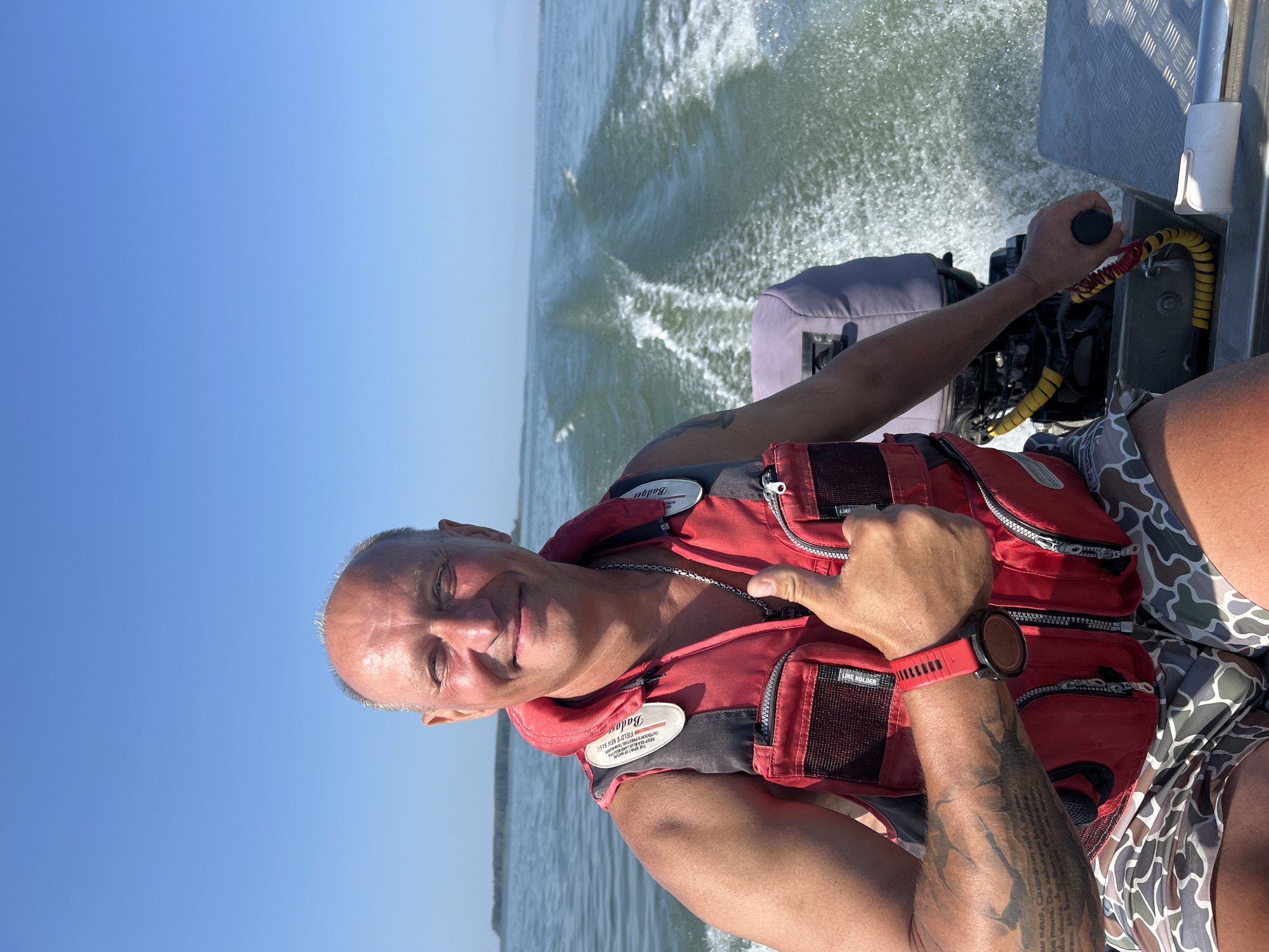 In the city of Beregovoe, Russia, a man with a shaved head and a smile is giving a thumbs-up while riding in a boat. He is wearing a red life vest and camouflage shorts. He has tattoos on his arm. The boat is moving through the water, creating a wake. The sky is blue and clear.