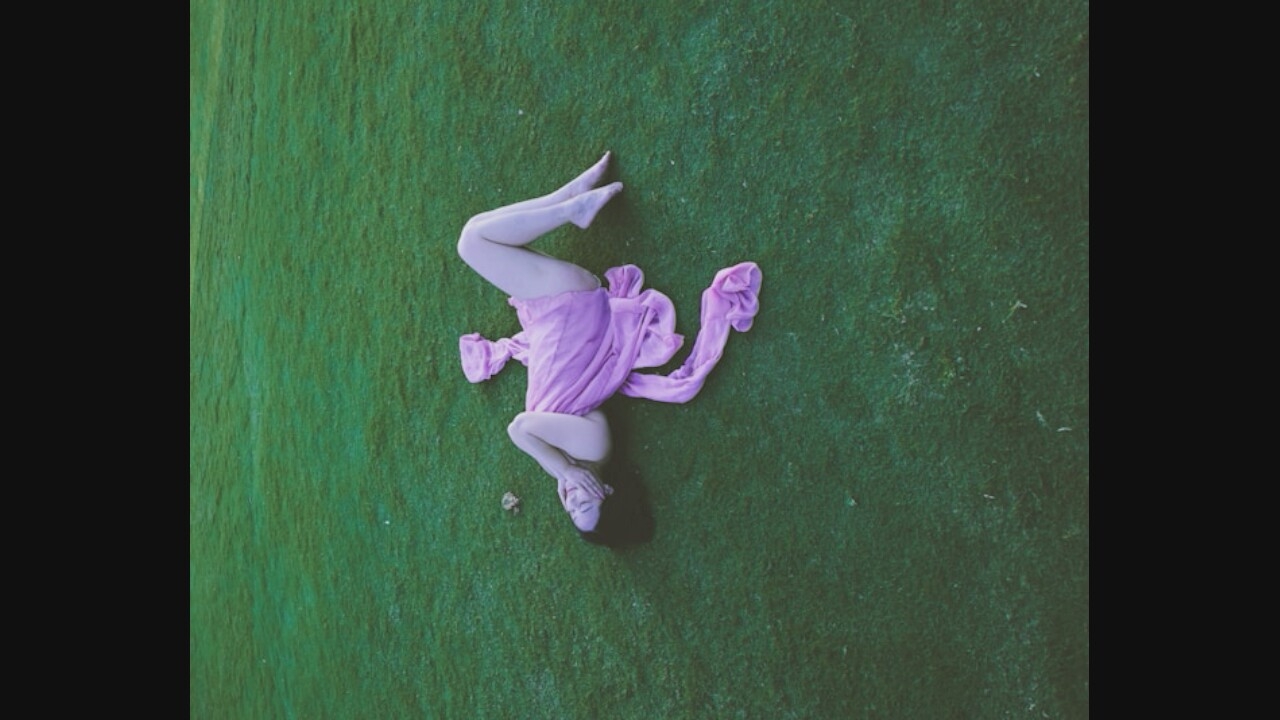 A person wearing a translucent purple fabric is lying on their back on a vibrant green grassy field. The fabric is draped around their body, with one end extending towards their head and the other trailing along the ground. The person's arms are bent, and their hands are near their face. Their legs are extended, with their feet pointed upwards. The setting appears to be outdoors, possibly a park or a well-maintained lawn. There are no visible structures or urban elements in the background, suggesting a natural or secluded environment.