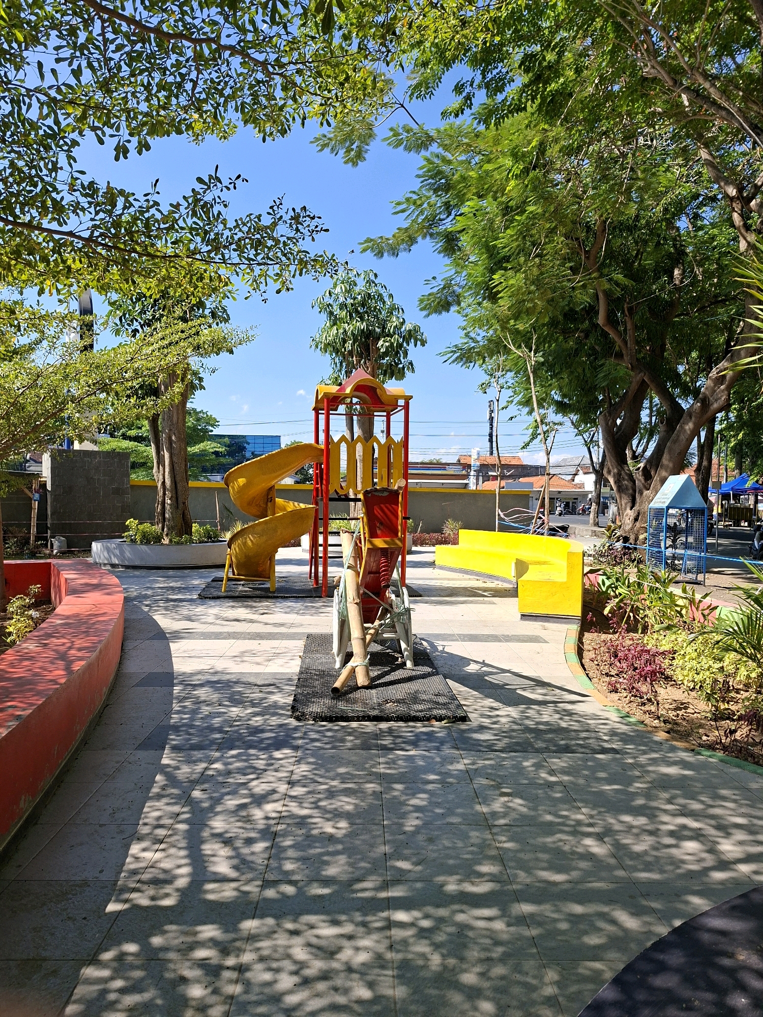 An outdoor playground is depicted with a yellow spiral slide, a red and yellow climbing structure, and a wooden see-saw. The playground is situated on a paved area with tree shadows cast across the ground. To the left, a red curved bench is visible, and to the right, a yellow bench is present along with landscaping featuring low-lying plants and foliage. In the background, trees and buildings suggest an urban setting. The sky is clear and blue. This scene is located in the city of Gunung Sekar, Indonesia.