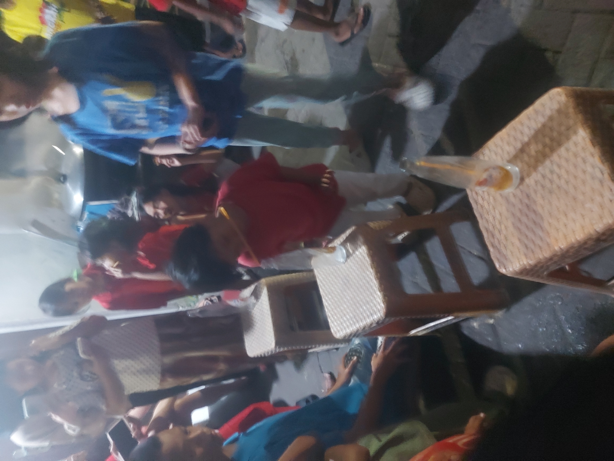 A group of people, mostly children and teenagers, are gathered outdoors, possibly in a residential area of Makassar, Indonesia. In the foreground, two light-colored woven plastic stools are visible, with a partially empty bottle of what appears to be a soft drink on the right-hand stool. Several individuals are standing or sitting around these stools. A young boy in a red shirt is leaning forward towards the stool on the left, appearing to be reaching for something. To his left, a person in a blue shirt is looking down. In the background, other individuals are milling about, some partially obscured. The environment appears to be paved, and the lighting suggests it is either dusk or nighttime.