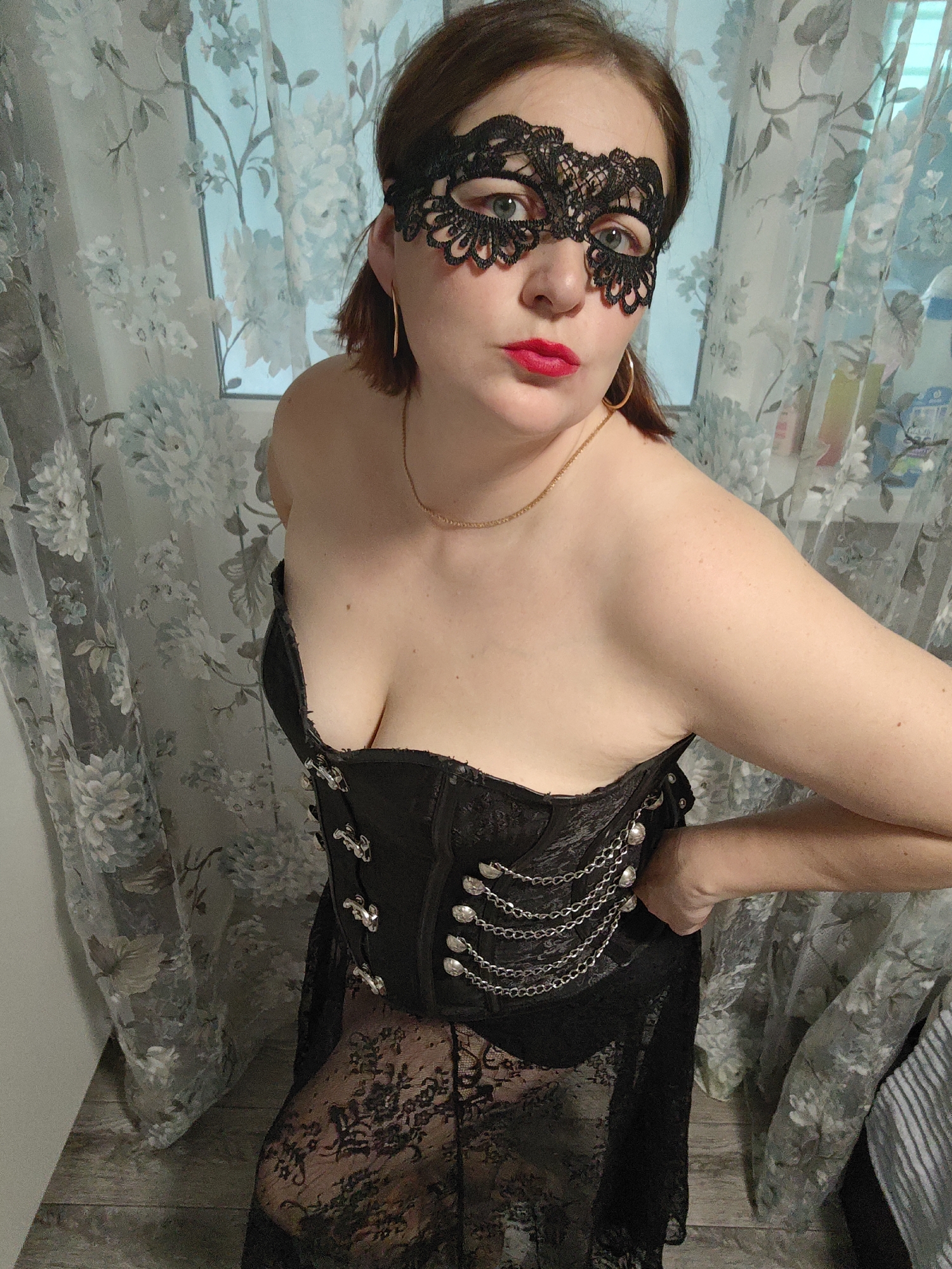 A woman is depicted in a portrait-style shot, looking directly at the camera. She has light brown hair, is wearing a black lace eye mask, and has bold red lipstick. She is also wearing large gold hoop earrings and a thin gold chain necklace. Her attire consists of a black strapless corset adorned with multiple silver chains and decorative buttons or clasps on the front, paired with a sheer black lace skirt or dress extending below the corset. Her arms are bent, with her hands on her hips, striking a confident pose. The background features light-colored curtains with a floral pattern in shades of blue, grey, and white, suggesting an indoor setting, possibly a residence in Markovskii, Russia. The lighting appears to be ambient, likely from a window behind the curtains, but the exact time of day or outdoor weather is not discernible. No visible text is present in the image.