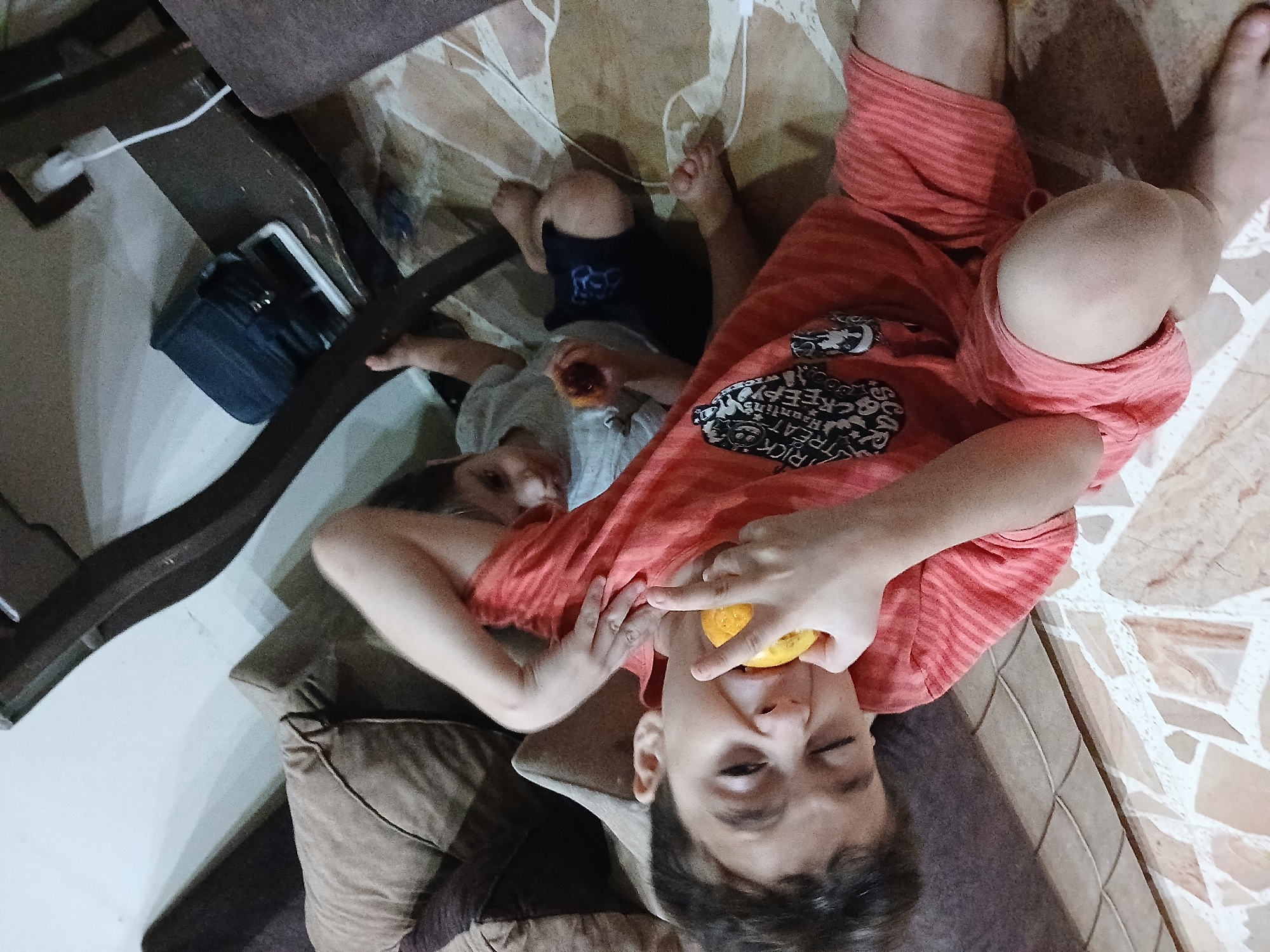 In Barza Municipality, Syria, two young boys are lying on the floor. The boy in the foreground, wearing a coral-colored t-shirt and shorts, has his eyes closed and is biting into a piece of orange. His left hand is near his mouth, and his right arm is bent at the elbow. Behind him, another boy in a light gray t-shirt and dark shorts is also lying down, looking towards the foreground boy. The floor is tiled with a beige and white patterned design. To the left of the boys are cushions from a sofa or bed. In the background, a dark wooden furniture piece is visible, with a white electrical outlet and a white cable plugged into it. A dark blue object, possibly a bag, rests on the furniture. The lighting suggests an indoor setting.