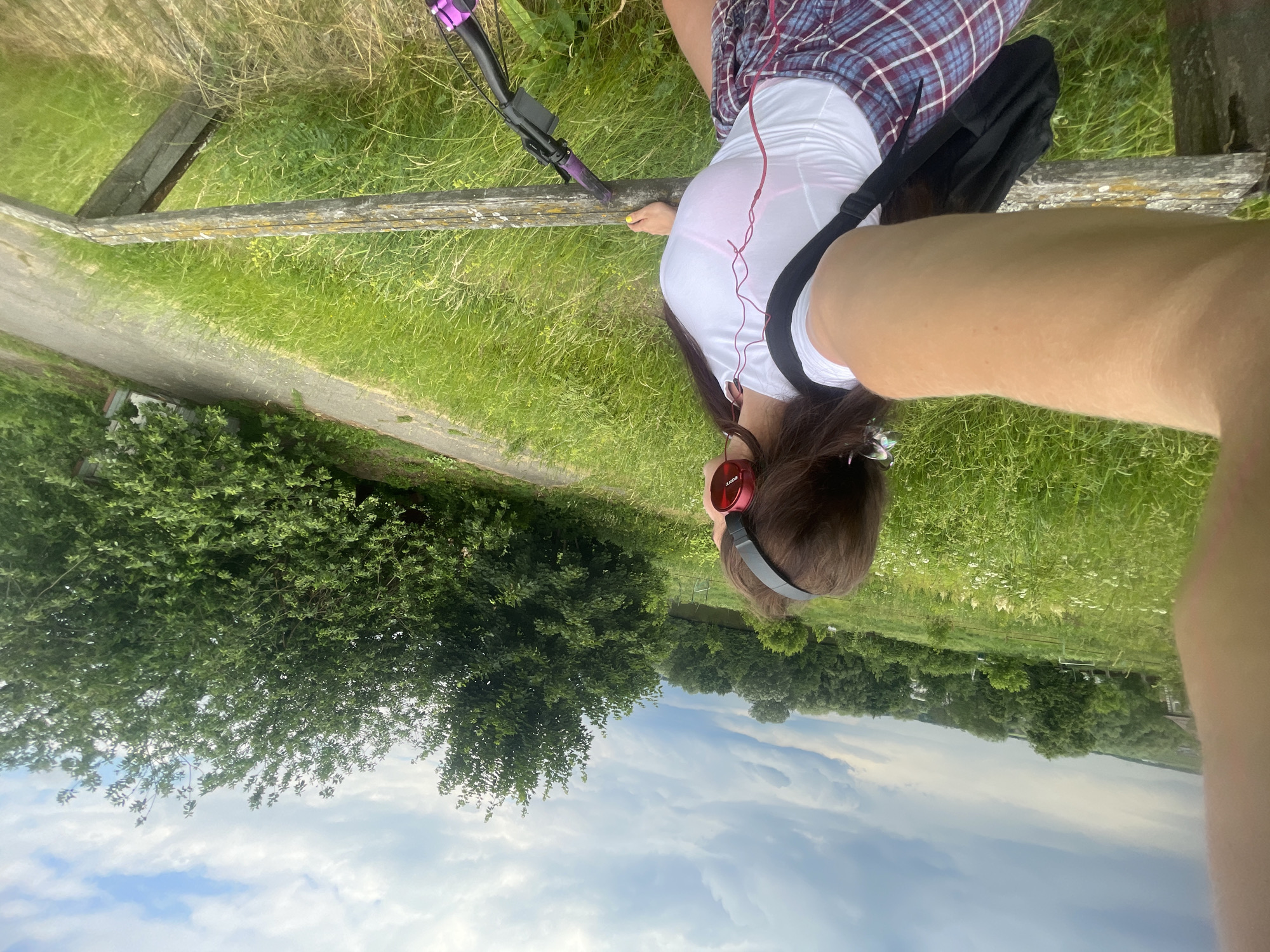 In Ostrov, Russia, on a cloudy day, a person stands on a wooden platform overlooking a grassy field with trees in the background. The person is wearing red headphones, a white t-shirt, and plaid shorts. A backpack is slung over their shoulder. A bicycle is parked next to them. The scene appears to be a scenic overlook, possibly near a park or nature reserve.