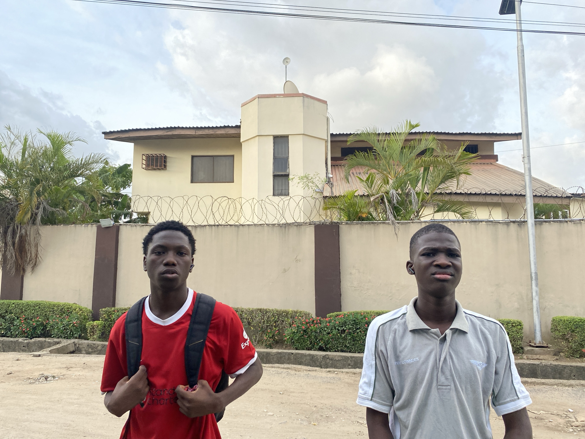 The image depicts two young men standing outdoors in what appears to be a residential area, consistent with a setting in Lagos, Nigeria. The sky is overcast with a blanket of clouds, suggesting a cloudy day or soft diffused light.
In the foreground, on the left, a young man with short dark hair is wearing a red t-shirt with white trim and a black backpack. Partial text on his shirt reads "Standard Chartered." He looks directly at the camera with a neutral expression. To his right, another young man, also with short dark hair, wears a light grey polo shirt with white stripes on the sleeves. The name "RYA LOWNDES" and a winged logo are visible on his shirt. He too looks at the camera with a neutral expression. Both men appear to have small earbuds in their left ears. The ground beneath them is unpaved, consisting of dirt or a rough pathway.
Behind the individuals, a cream-colored perimeter wall topped with prominent razor wire stretches across the frame, indicating security measures. A well-maintained green hedge with small red flowers runs along the base of this wall. Behind the wall stands a two-story building, also cream-colored, featuring a distinctive central octagonal tower section with narrow windows. A satellite dish and a small antenna are visible on the roof of this tower. The building has a corrugated metal roof and multiple windows, some fitted with security grates. Lush green palm trees and other tropical foliage are visible around and behind the building. Overhead, utility wires cross the sky, and a tall light pole, possibly with a solar panel, stands on the far right.